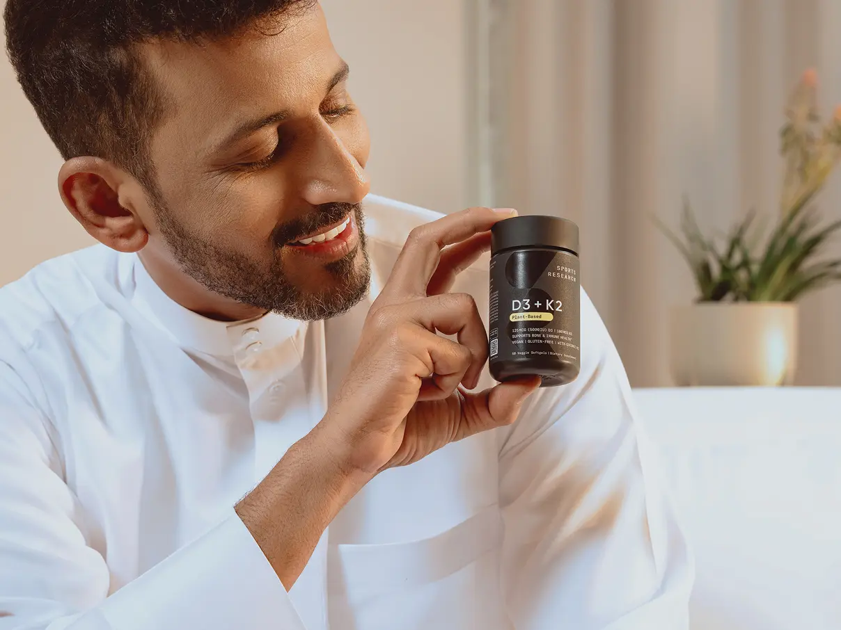 iHerb logo. In the background a smiling man sits on a white sofa, wearing traditional Arabic clothing and holding an iHerb product.