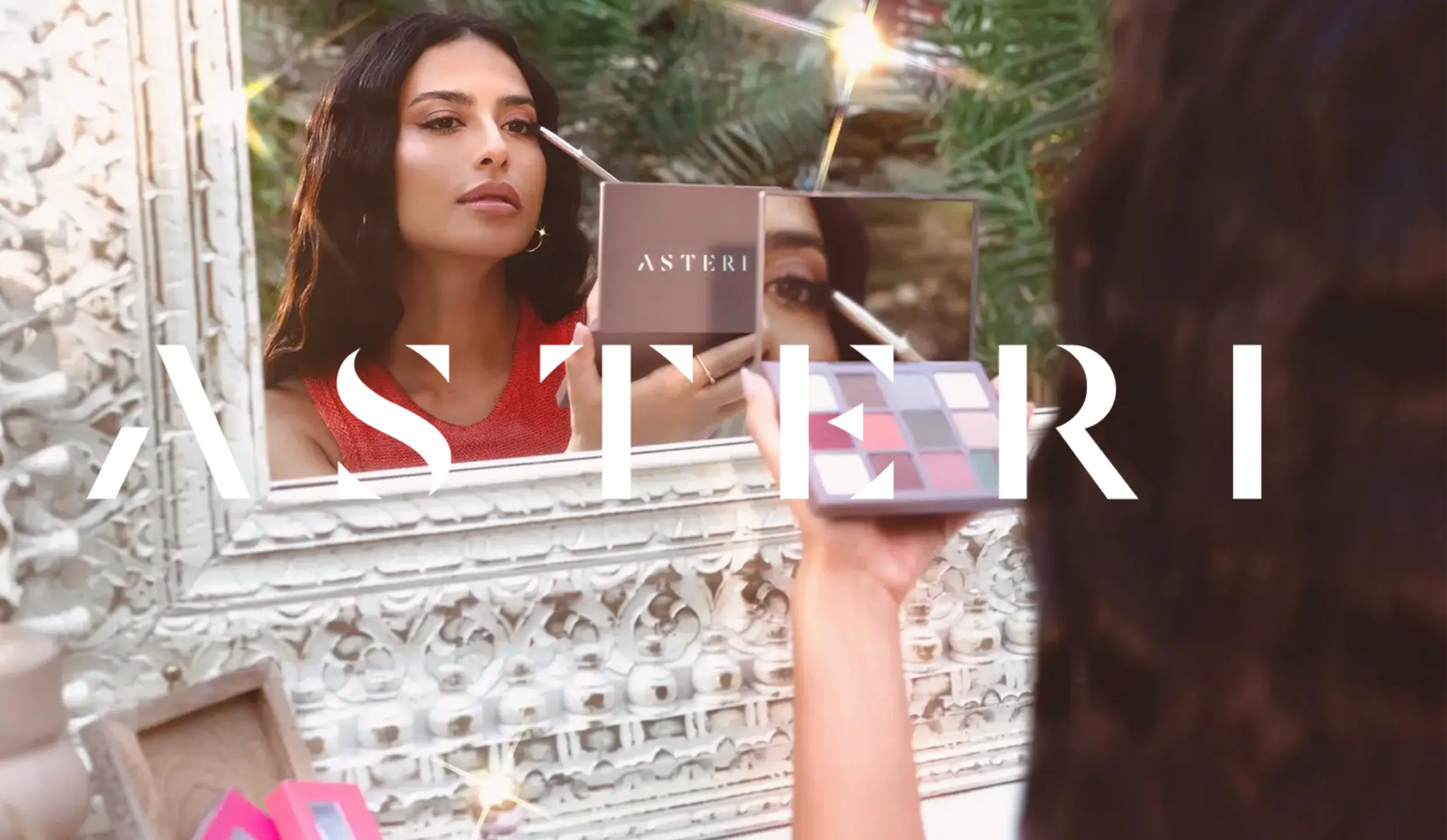 Asteri logo. In the background a woman is standing in front of a mirror, applying mascara.