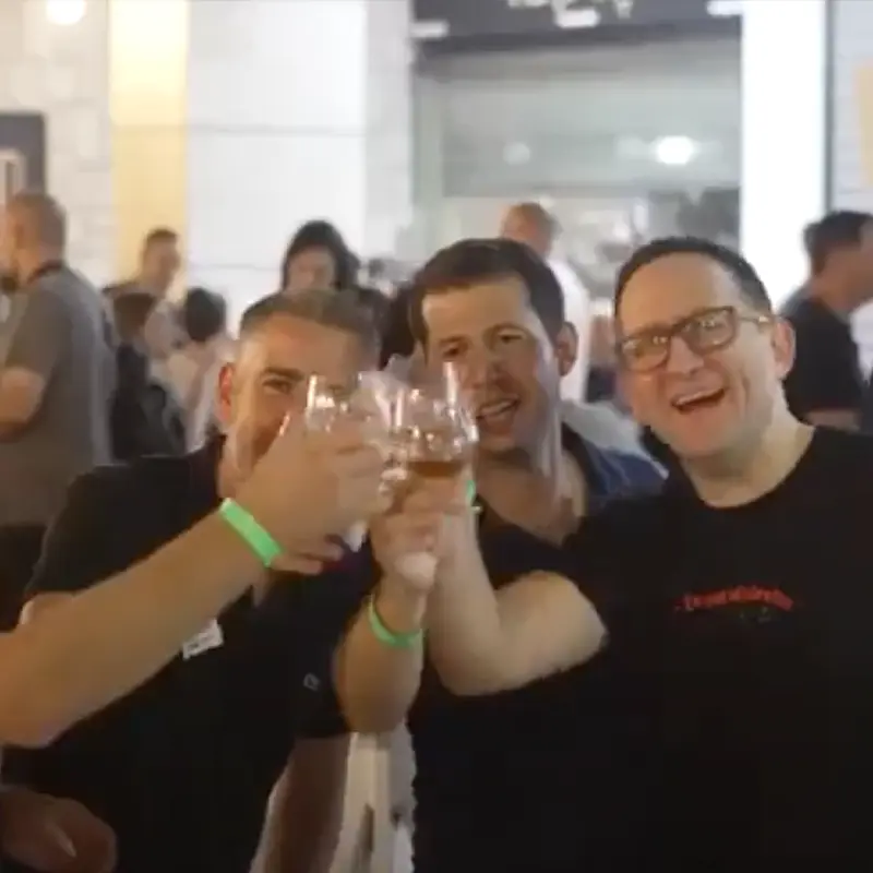 Three men smiling and clinking glasses in a celebratory toast at an outdoor gathering.