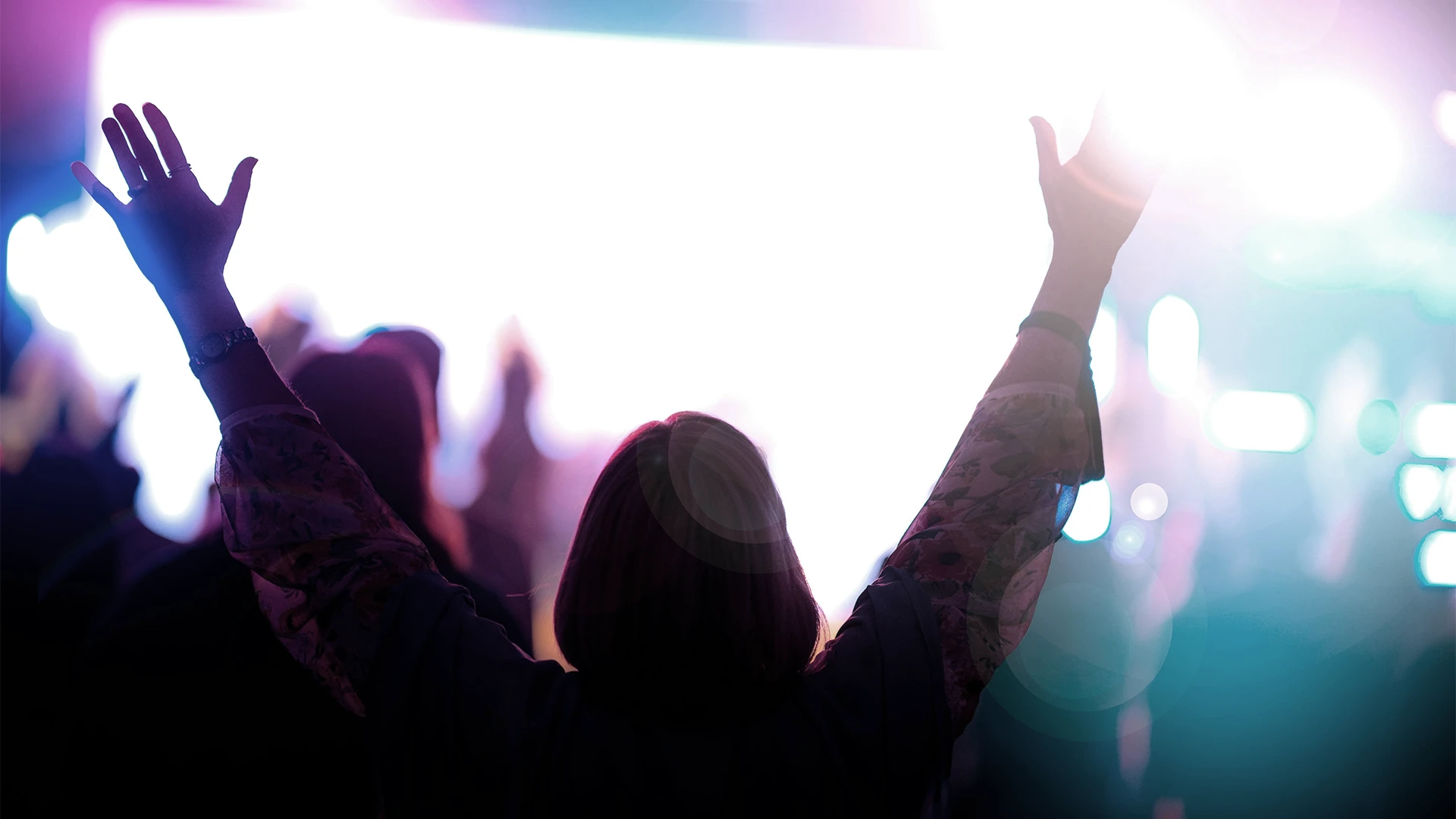 Person with raised hands in a crowd, lit by bright colorful stage lights.