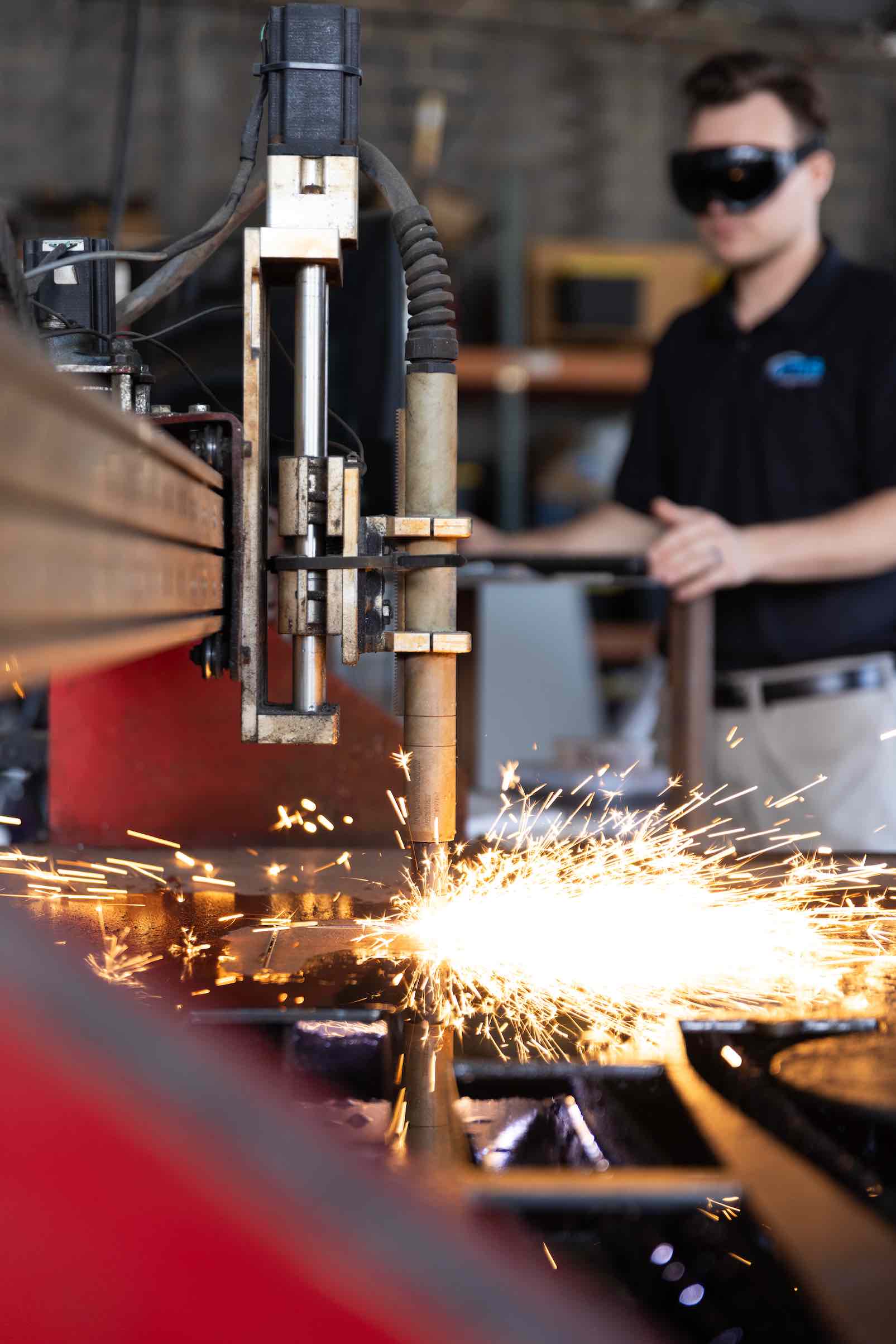 A welding or cutting machine emits bright sparks during operation, while a person in protective goggles stands blurred in the background, observing the process