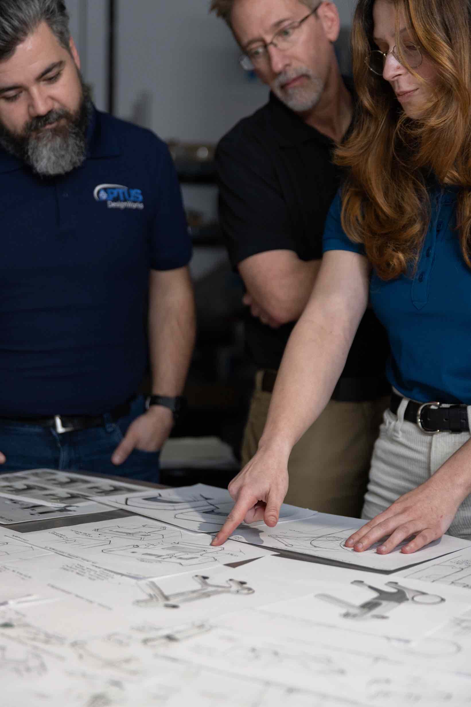 3 people in APTUS polo shirts collaborating on product designs, looking at paper sketches on a table with the person on the right pointing to a specific piece of paper