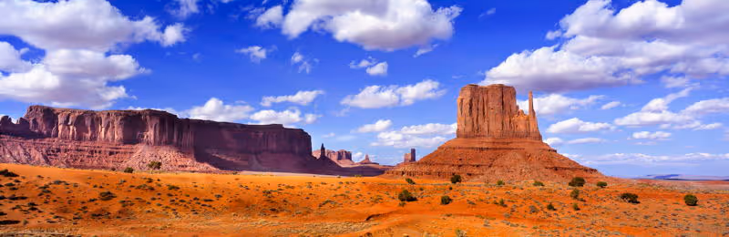 Wide desert landscape with red rock formations under a bright blue sky with scattered white clouds.