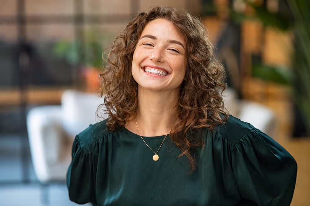 Smiling woman with curly brown hair wearing a dark green blouse and a gold pendant necklace.