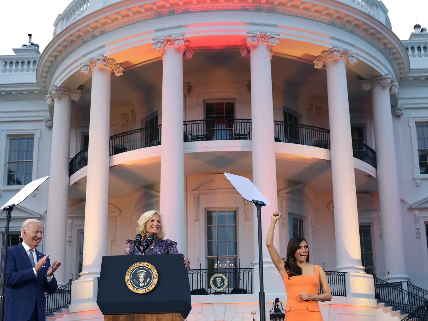 Eva Longoria in orange dress raising her fist in celebration as First Lady Jill Biden and President Joe Biden applaud at formal White House event