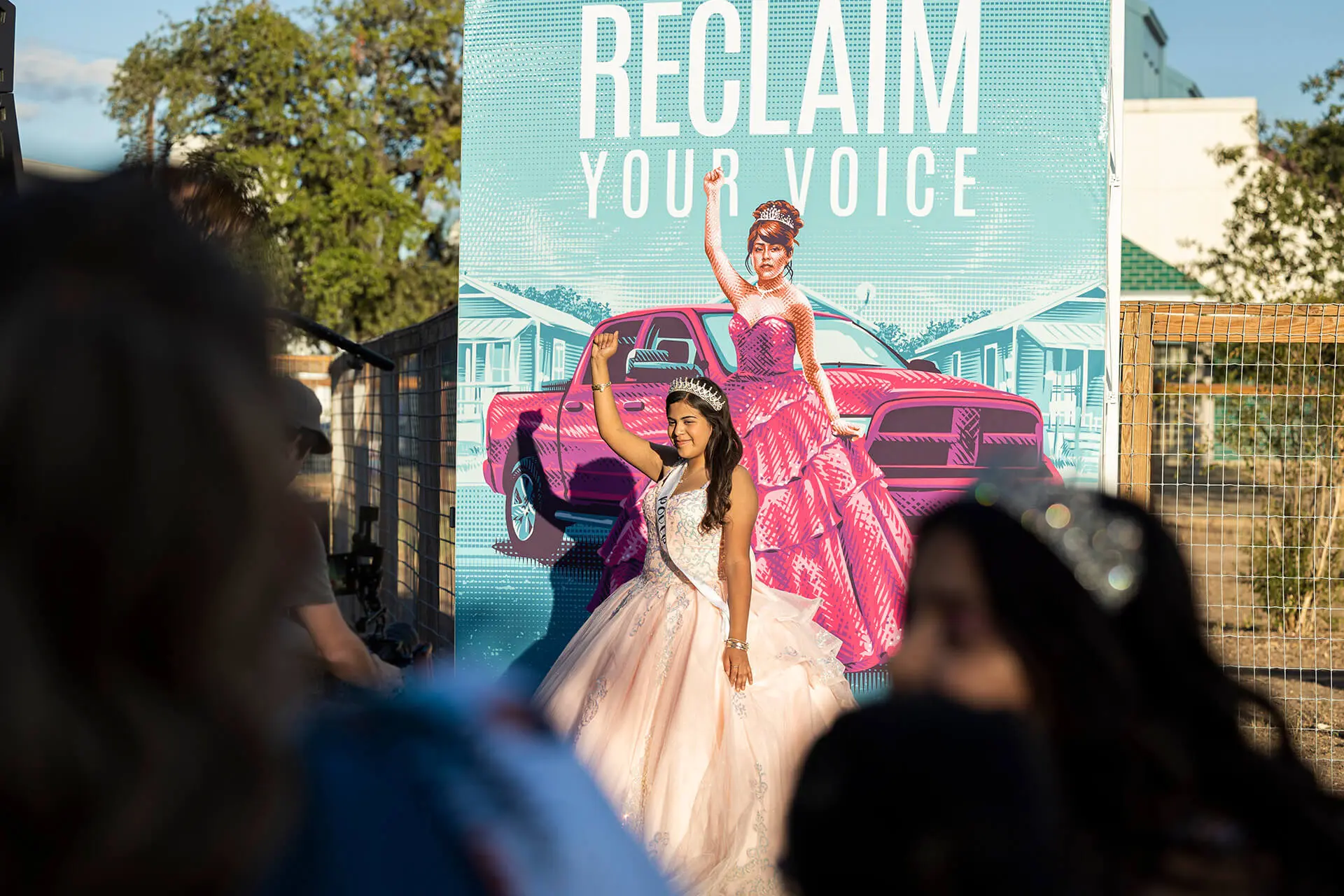 Young woman in white quinceañera dress and tiara posing in front of 'Reclaim Your Voice' mural with pink truck at Quince to the Polls voter engagement event