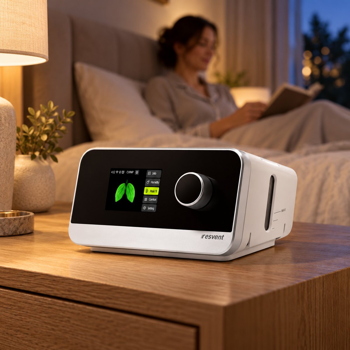 Equipo CPAP Resvent iBreeze sobre la mesa de noche al anochecer mientras una mujer lee en la cama