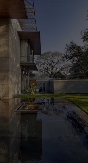 Modern house with large windows beside a reflecting pool and a green lawn bordered by a concrete wall with trees in the background.