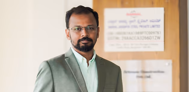 Man with glasses and beard wearing a light green suit jacket and white shirt standing indoors.