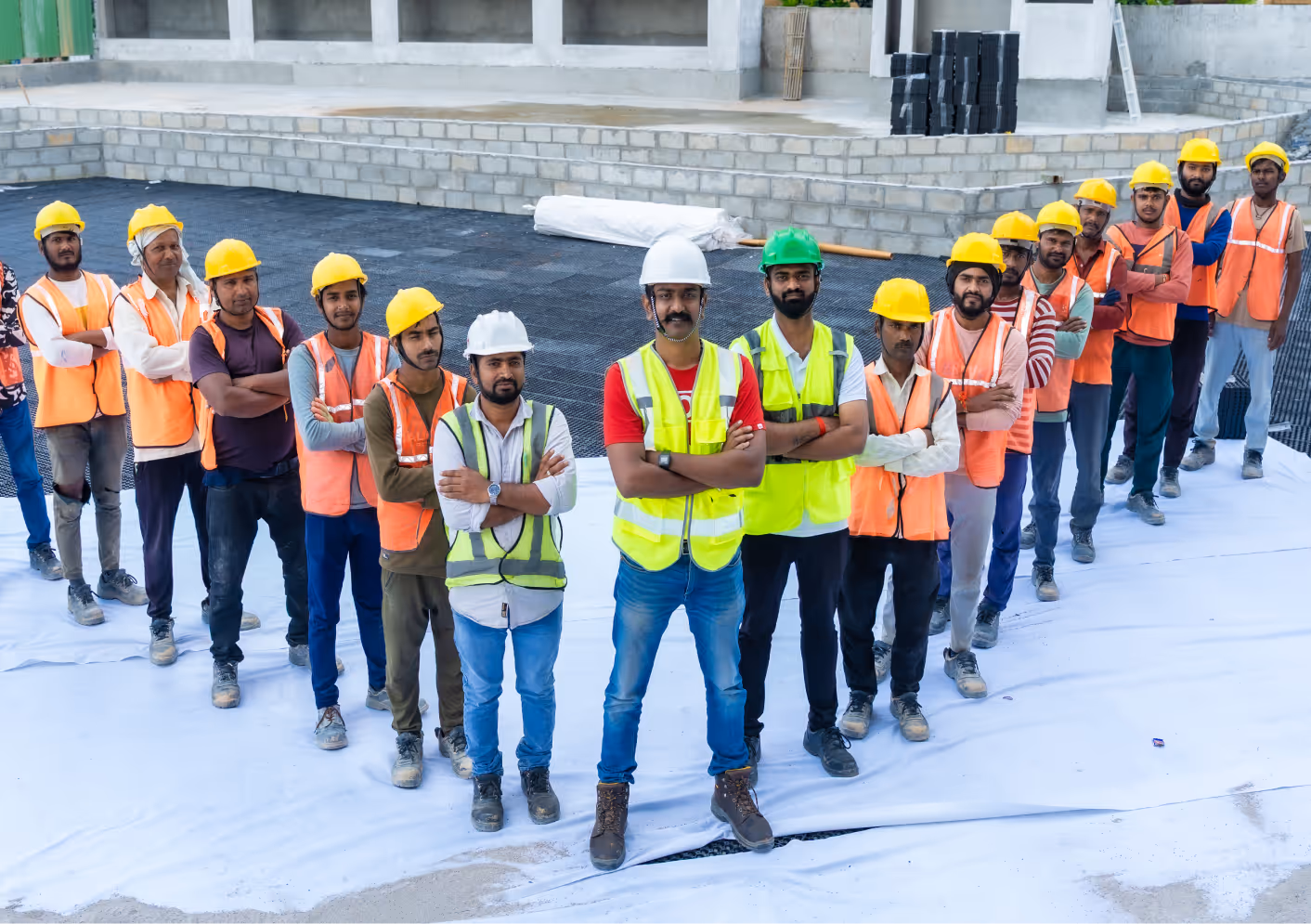 Group of construction workers wearing safety helmets and vests standing on a construction site with arms crossed.