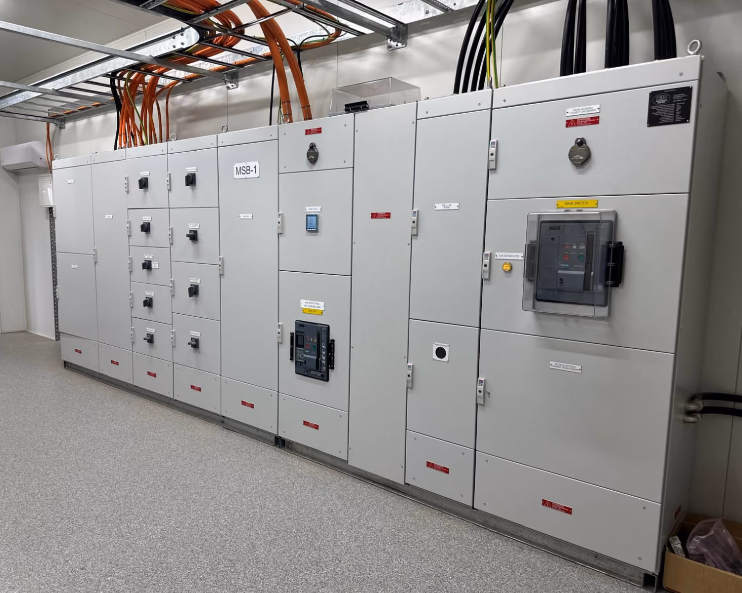Large gray industrial electrical switchboard with multiple panels and disconnected cables overhead.