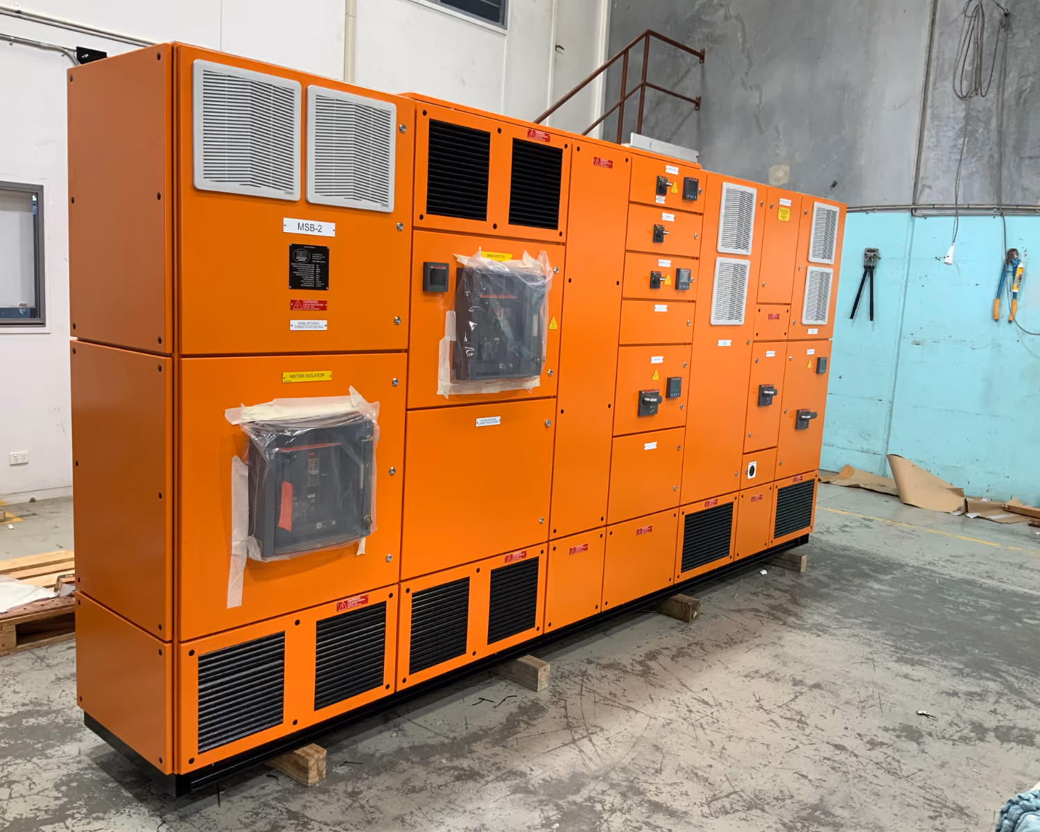 Long orange electrical switchboard unit inside industrial room with concrete floor and tools on wall.