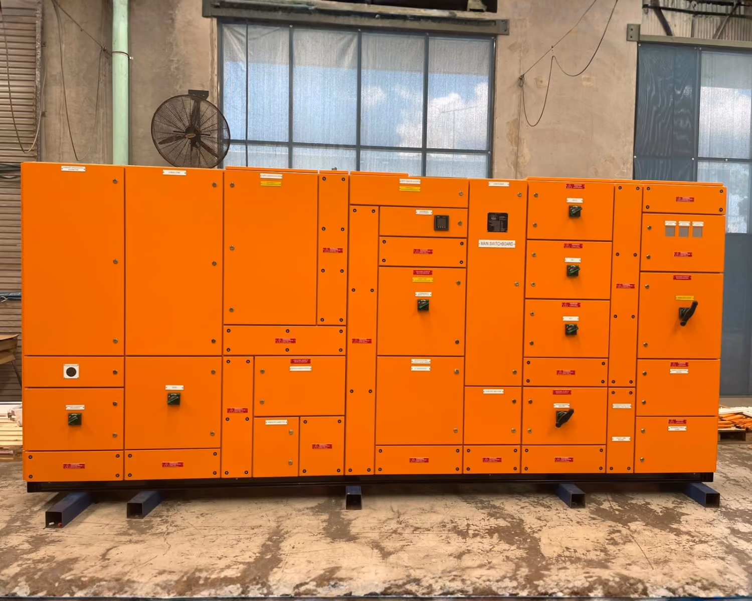 Large orange industrial switchboard with multiple panels and controls in warehouse setting.