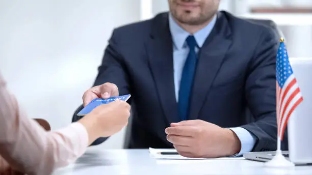 A person handing a document to a professional with a small U.S. flag on the desk. Beyond Border.