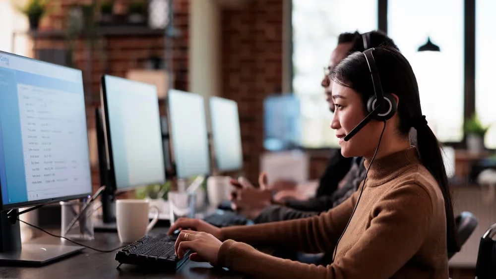 A customer service representative working at a desk with multiple monitors and a headset. Beyond Border.