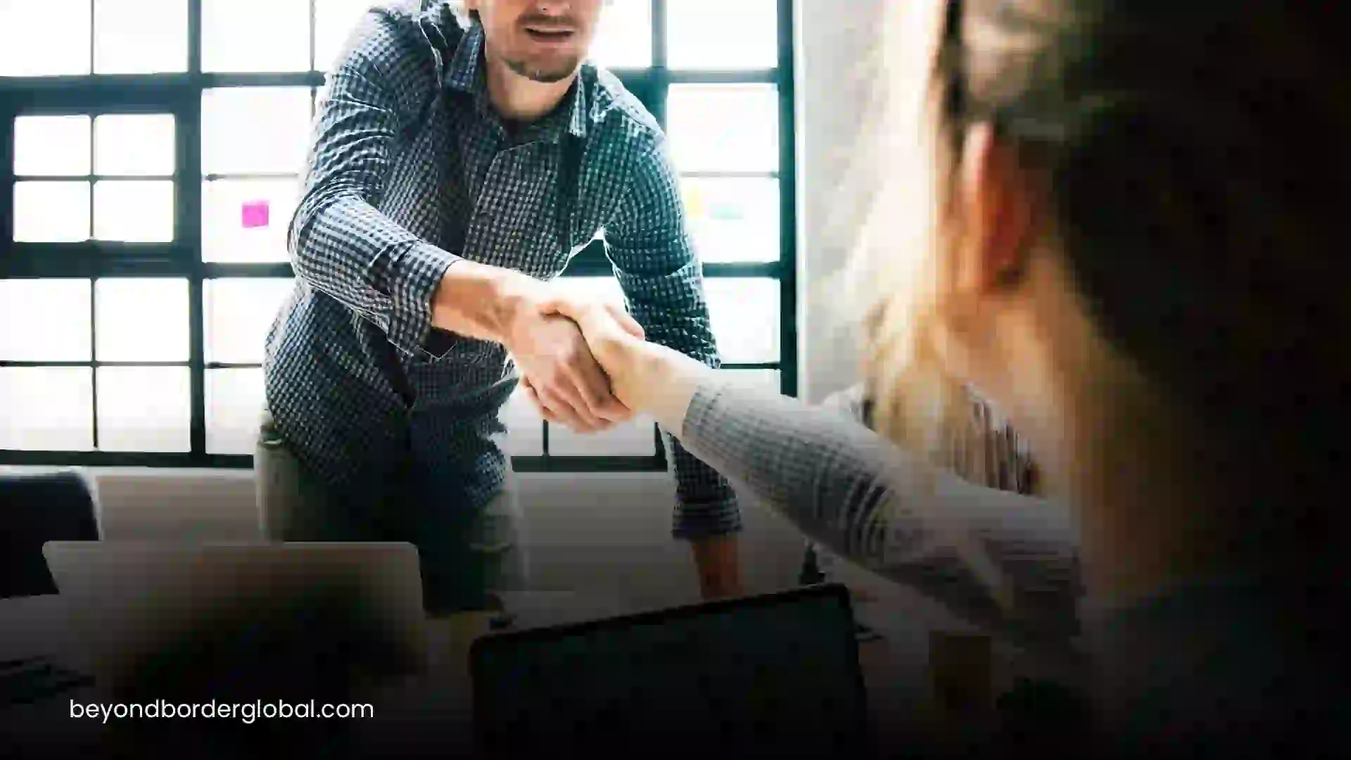 Two professionals shaking hands across a desk in an office. Beyond Border.