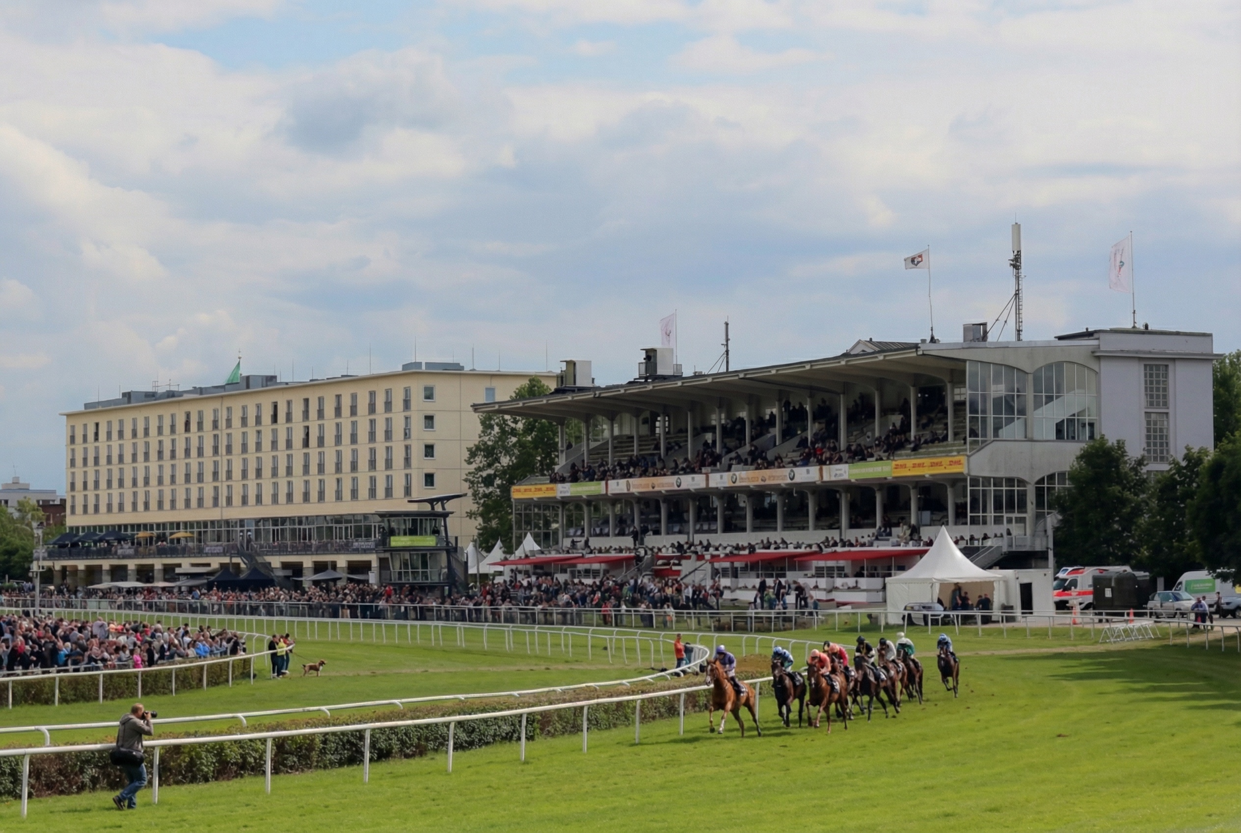 Pferderennen auf einer Grasrennbahn mit Zuschauern in einem mehrstöckigen Tribünengebäude und einem großen Hotel im Hintergrund Horn, Hamburg.