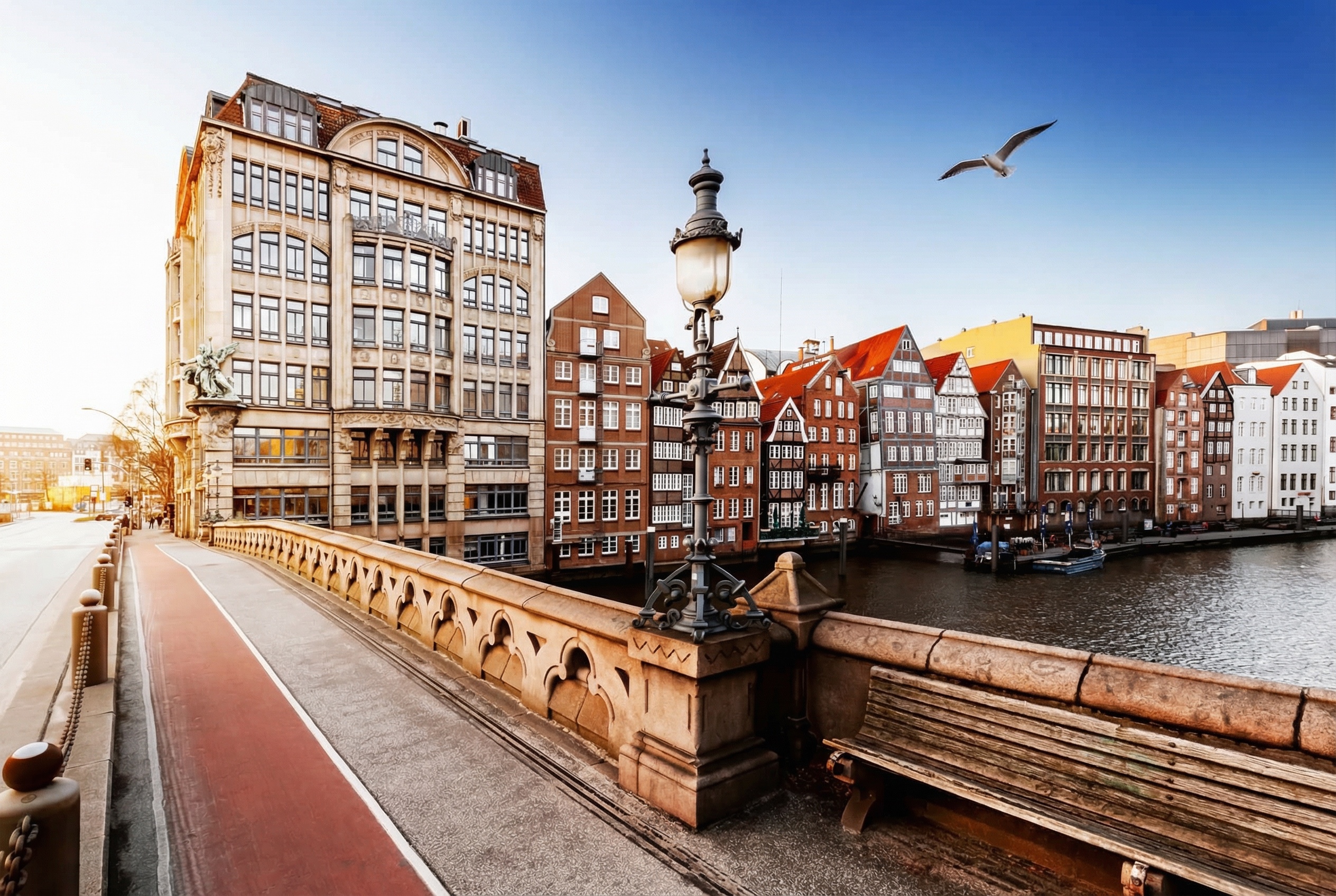 Blick auf eine Brücke, Nikolaifleet Hamburg, mit historischer Straßenlaterne, Sitzbank und Backsteinhäusern am Wasser unter klarem Himmel.