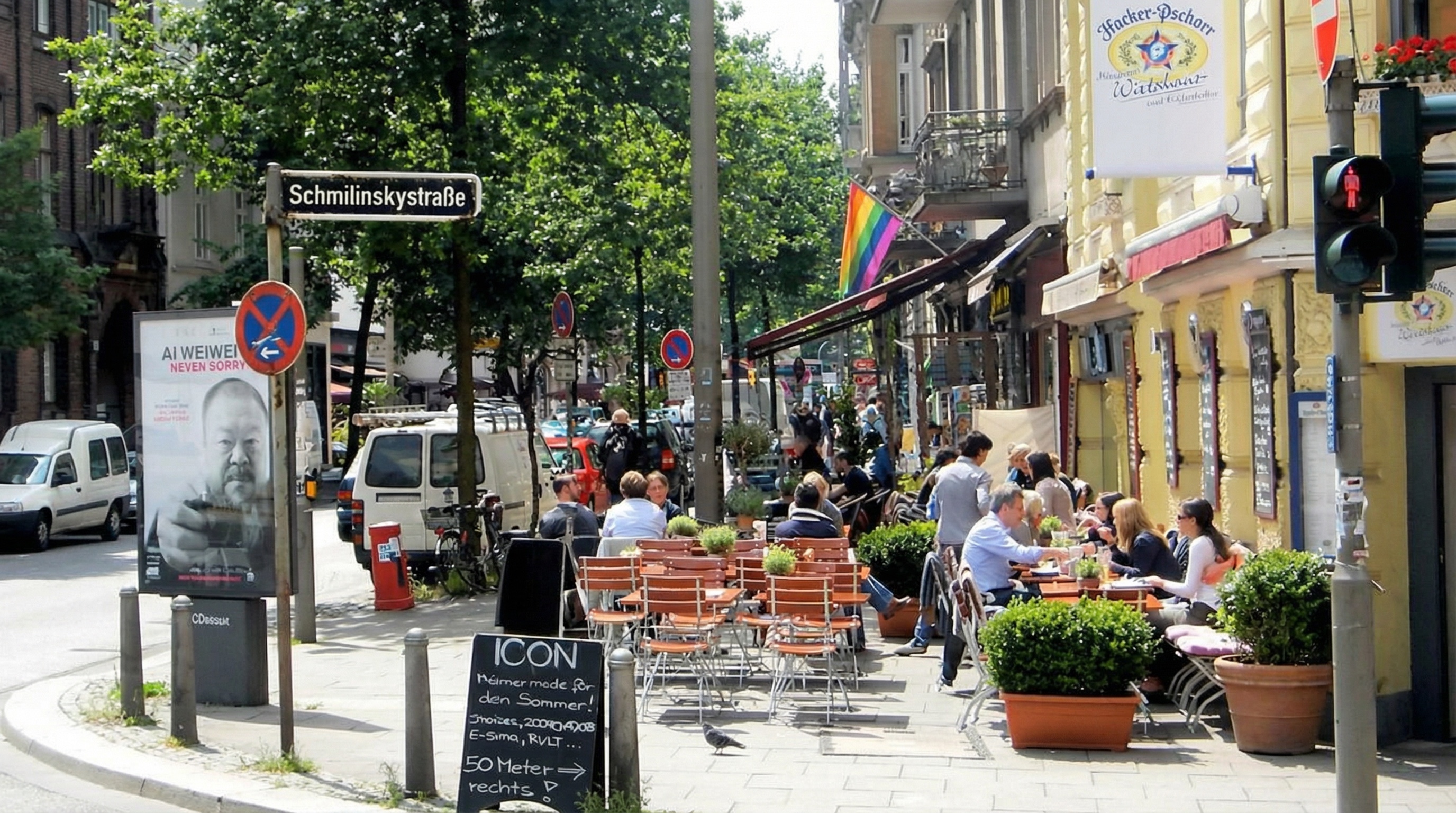 Straßenszene mit vielen Menschen, die an Außentischen eines Cafés sitzen, umgeben von Pflanzen und Straßenschildern in St. Georg, Hamburg.