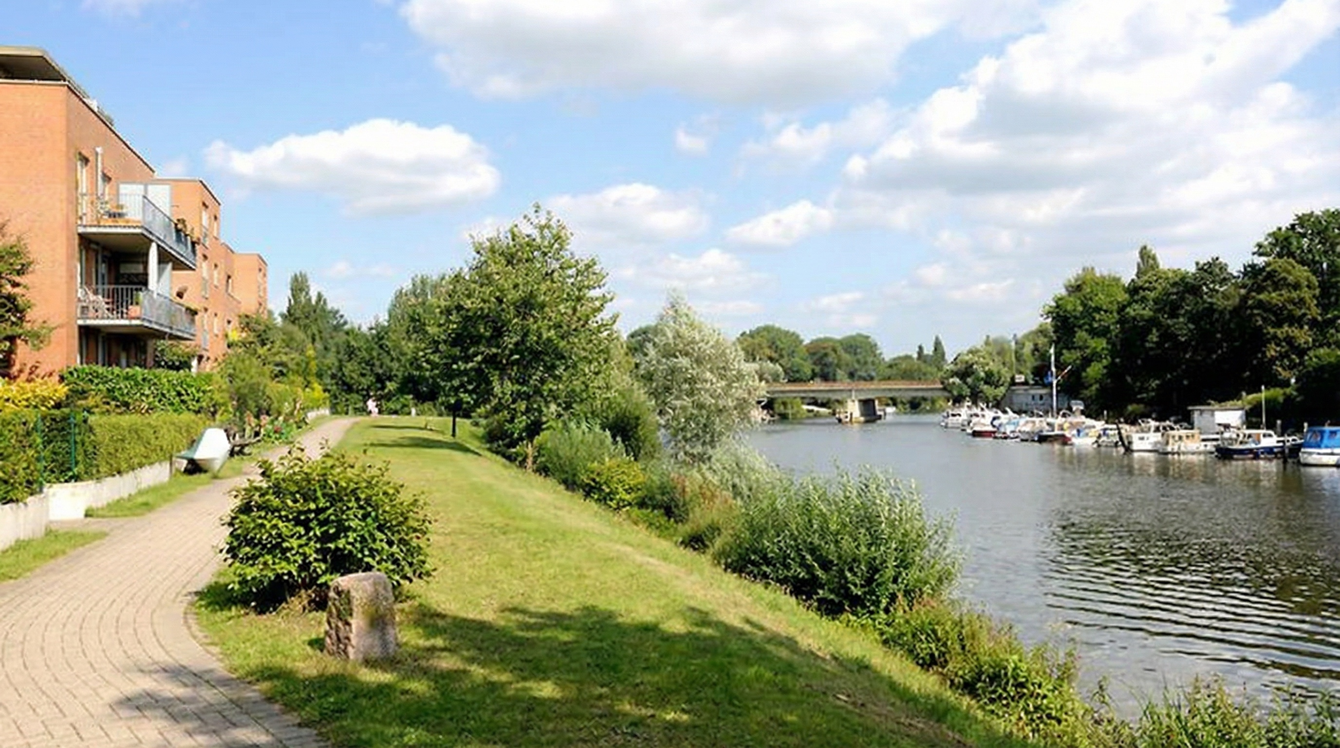 Grüner Uferweg mit Büschen und Bäumen neben einem Fluss, auf dem mehrere Boote liegen, unter blauem Himmel mit Wolken in Hamm, Hamburg.