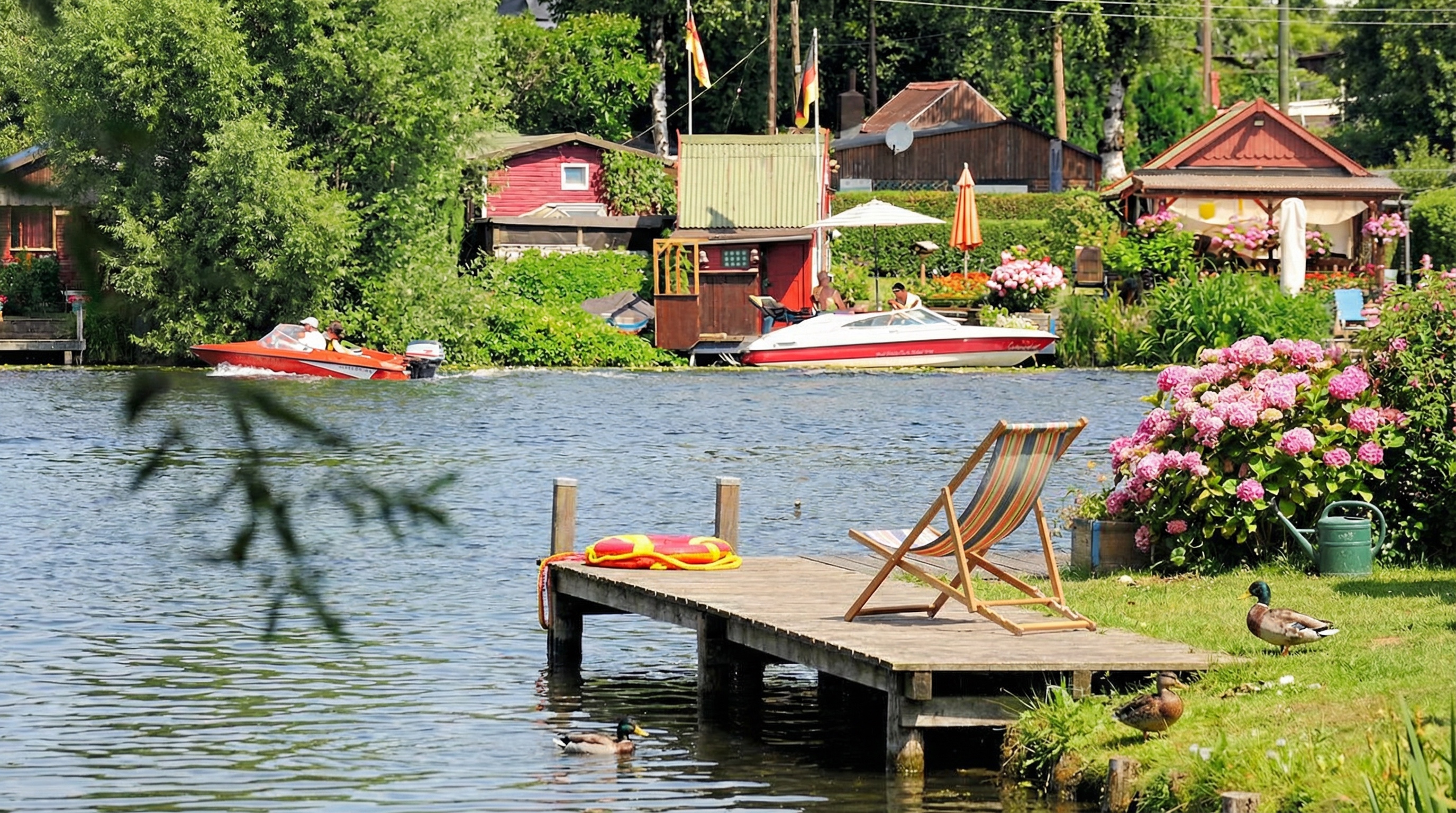 Liegestuhl auf Holzsteg am Ufer mit Blumen, Enten und Booten auf dem Wasser in grüner Umgebung in Rothenburgsort, Hamburg.