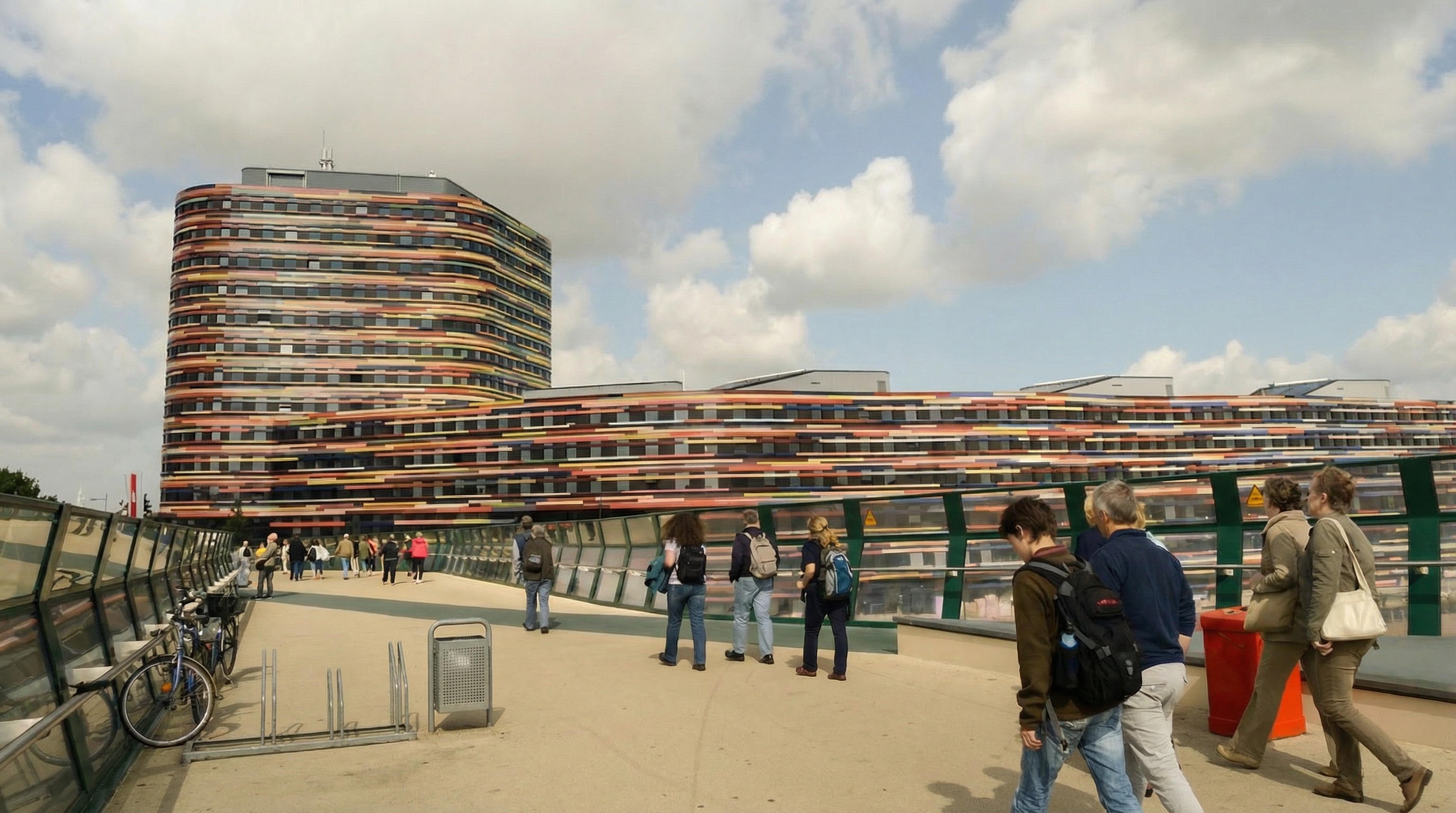 Mehrstöckiges, langgestrecktes Gebäude mit abgerundeter Ecke und horizontalen, farbigen Streifen, davor Fußgänger auf breitem Gehweg in Wilhelmsburg, Hamburg.