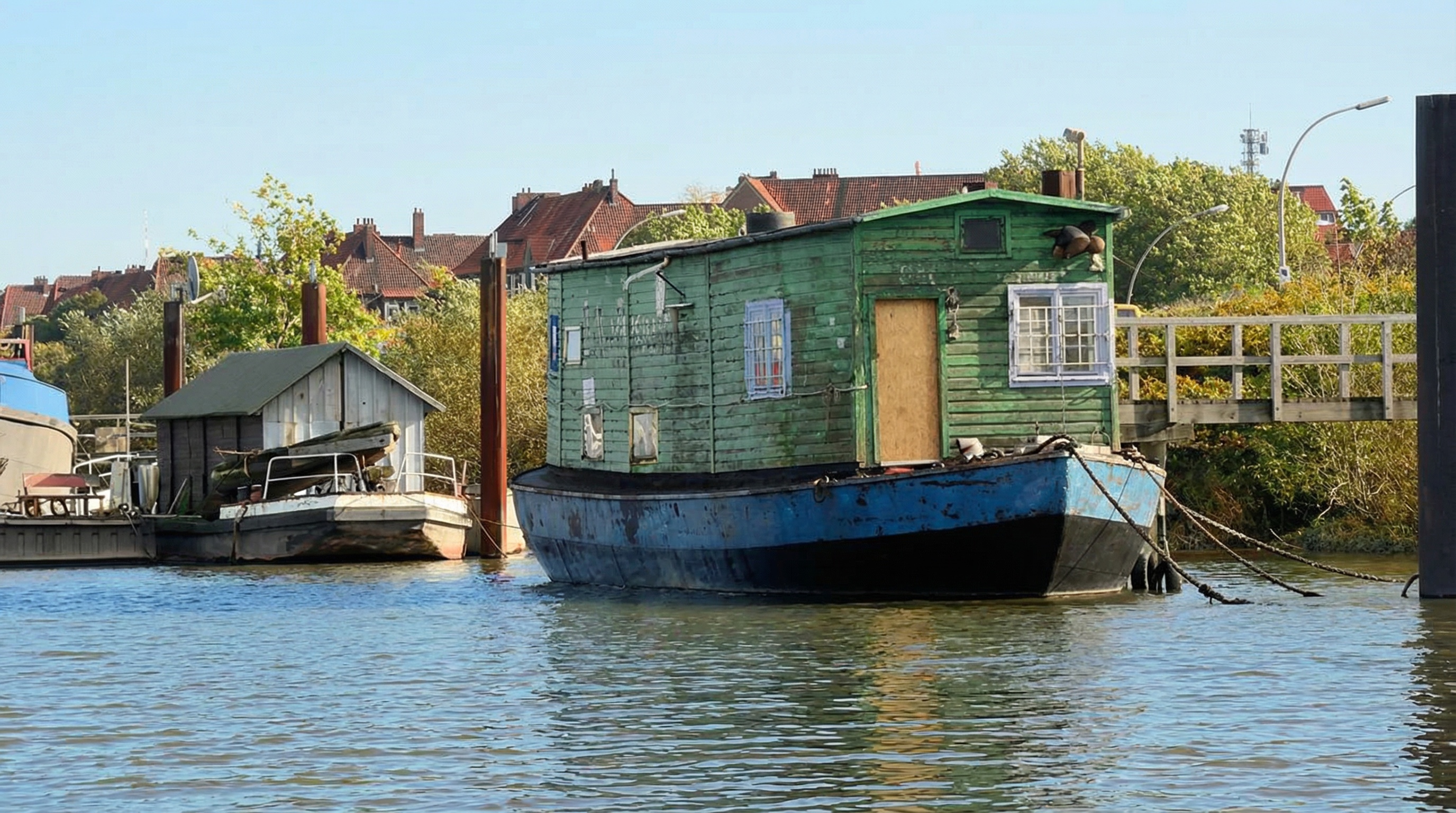 Grünes Hausboot mit blauer Unterseite liegt am Ufer des Spreehafens, im Hintergrund Häuser und Bäume. Kleiner Grasbrook, Hamburg.