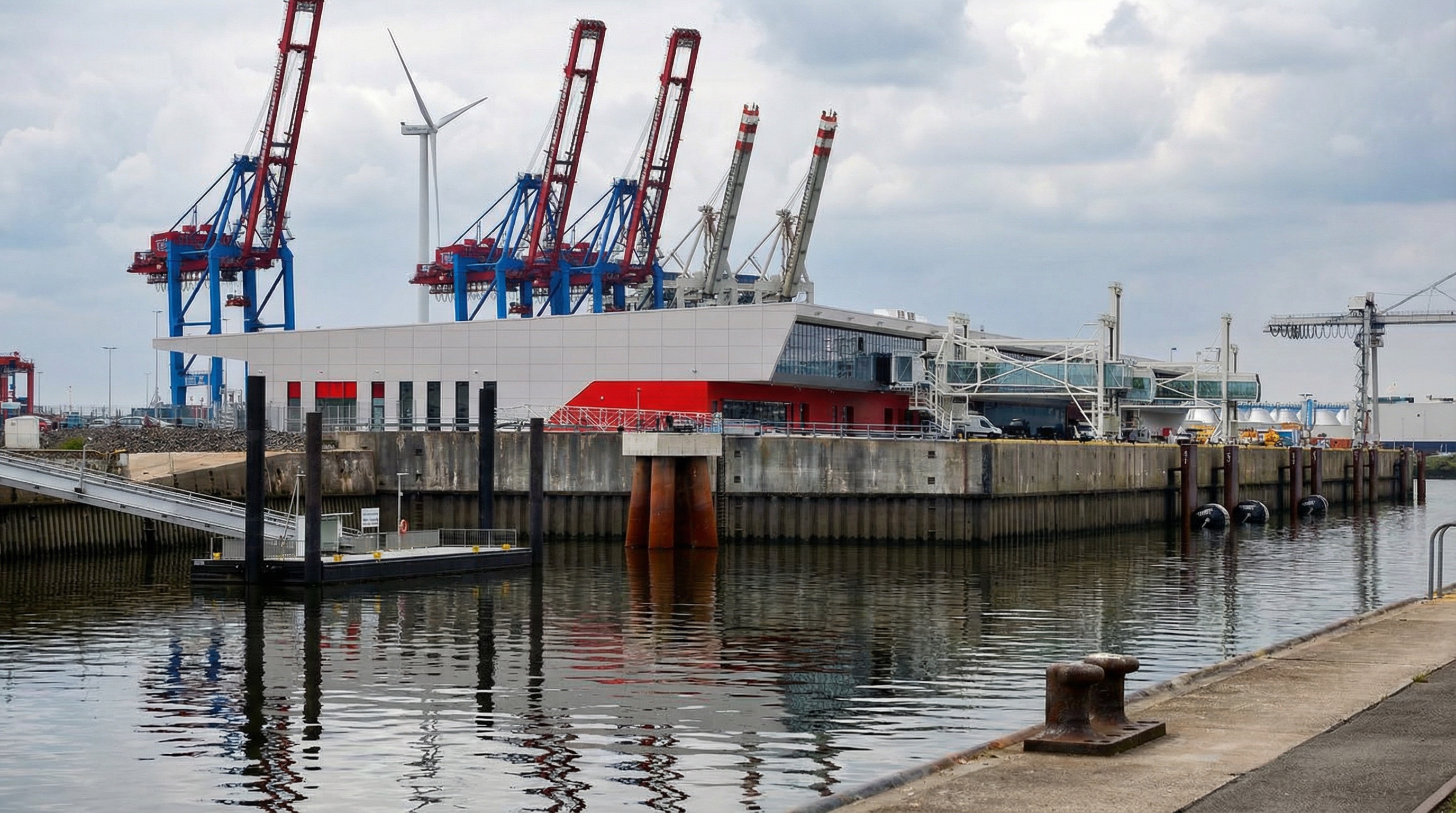 Hafenanlage des Hamburg Cruise Center mit mehreren großen Hafenkränen, einem Windrad im Hintergrund und einem Gebäude mit rotem und weißem Anstrich am Wasser in Steinwerder, Hamburg.