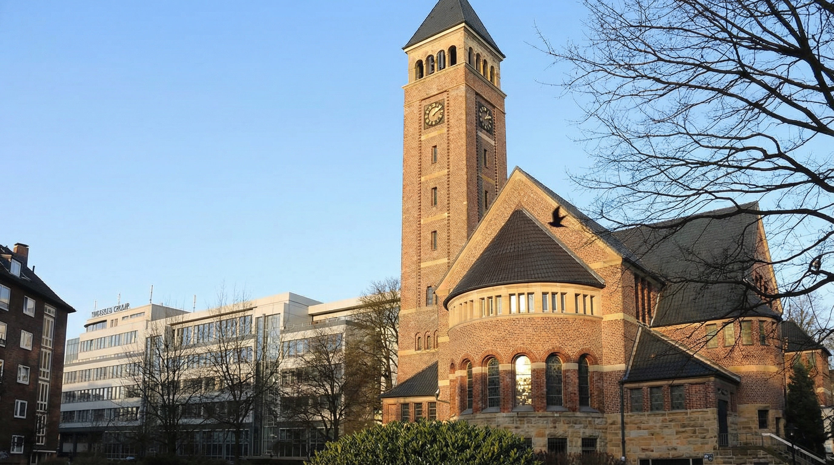 Kirchturm mit Uhr an einem Backsteingebäude, umgeben von Bäumen und modernen Gebäuden im Hintergrund in Altona-Nord, Hamburg.