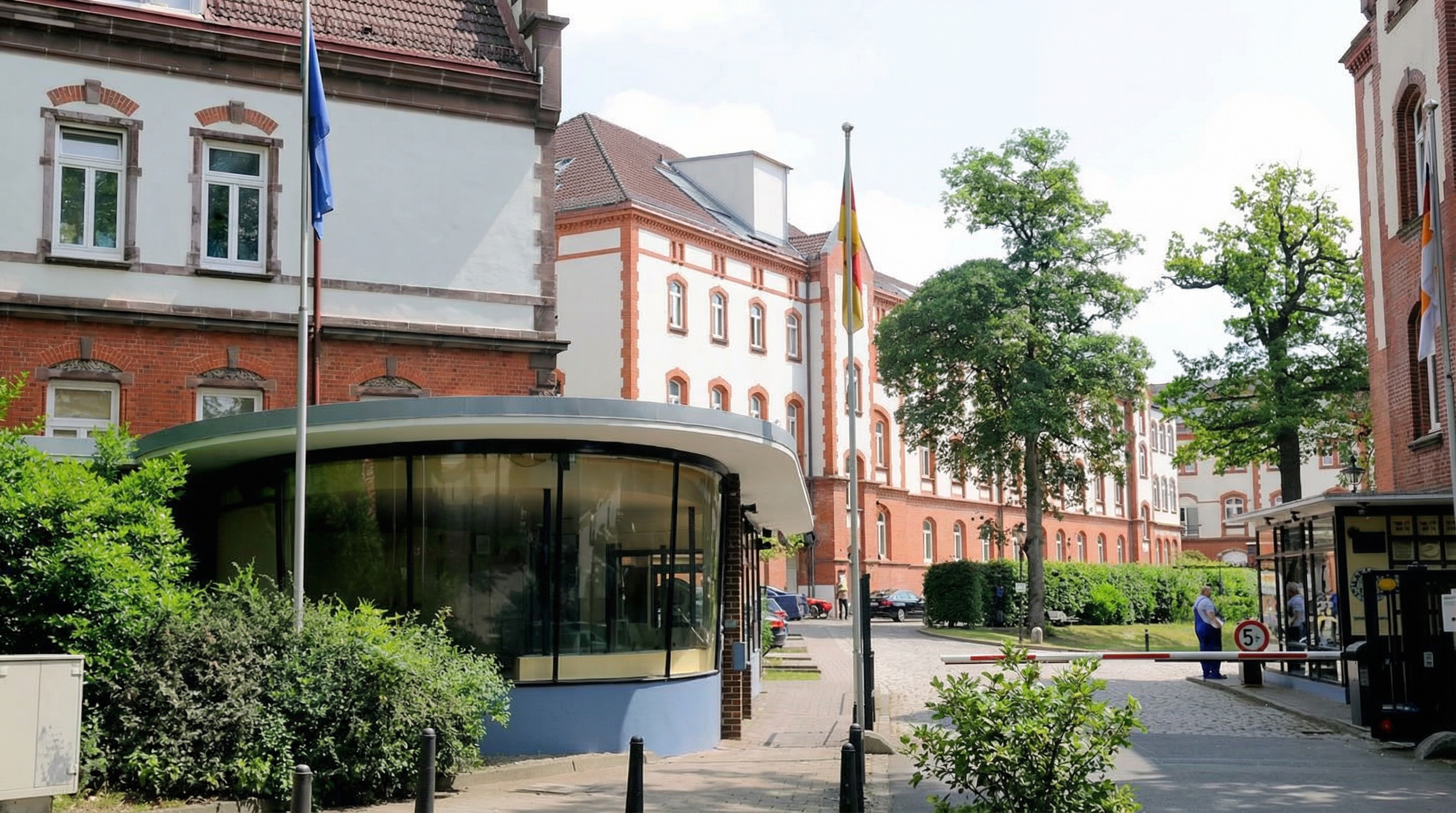 Gebäude mit rotem Backstein und weißen Fassaden, davor runde Glasstruktur, mehrere Fahnenmasten und Bäume auf einem gepflasterten Platz in Bahrenfeld, Hamburg.