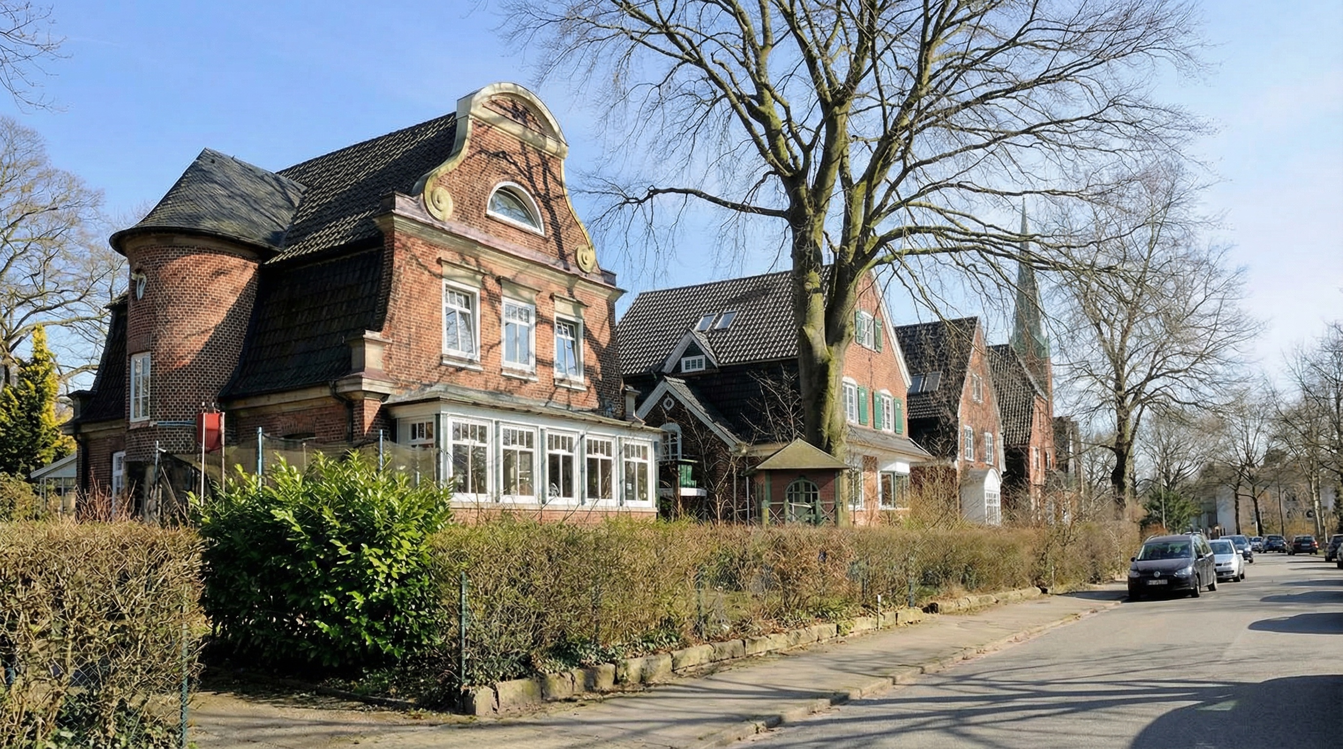 Reihe historischer Backsteinhäuser mit Giebeln und Gauben an einer ruhigen Straße, im Vordergrund ein kahler Baum und Hecke in Groß Flottbek, Hamburg.