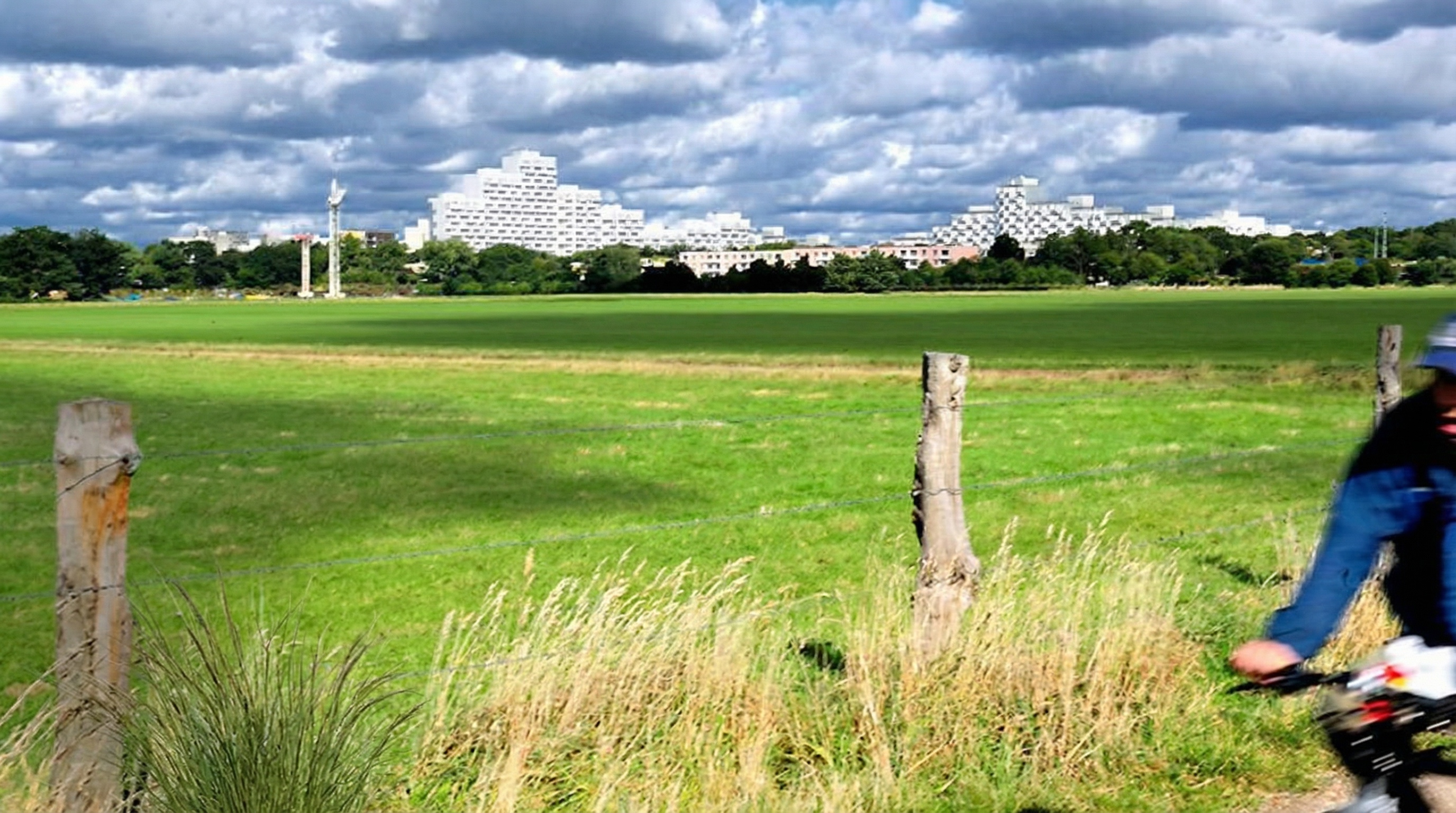 Grüne Wiese mit Holzzaunpfählen im Vordergrund, weiße Hochhäuser unter bewölktem Himmel, rechts Radfahrer im blauen Oberteil in Iserbrook, Hamburg.