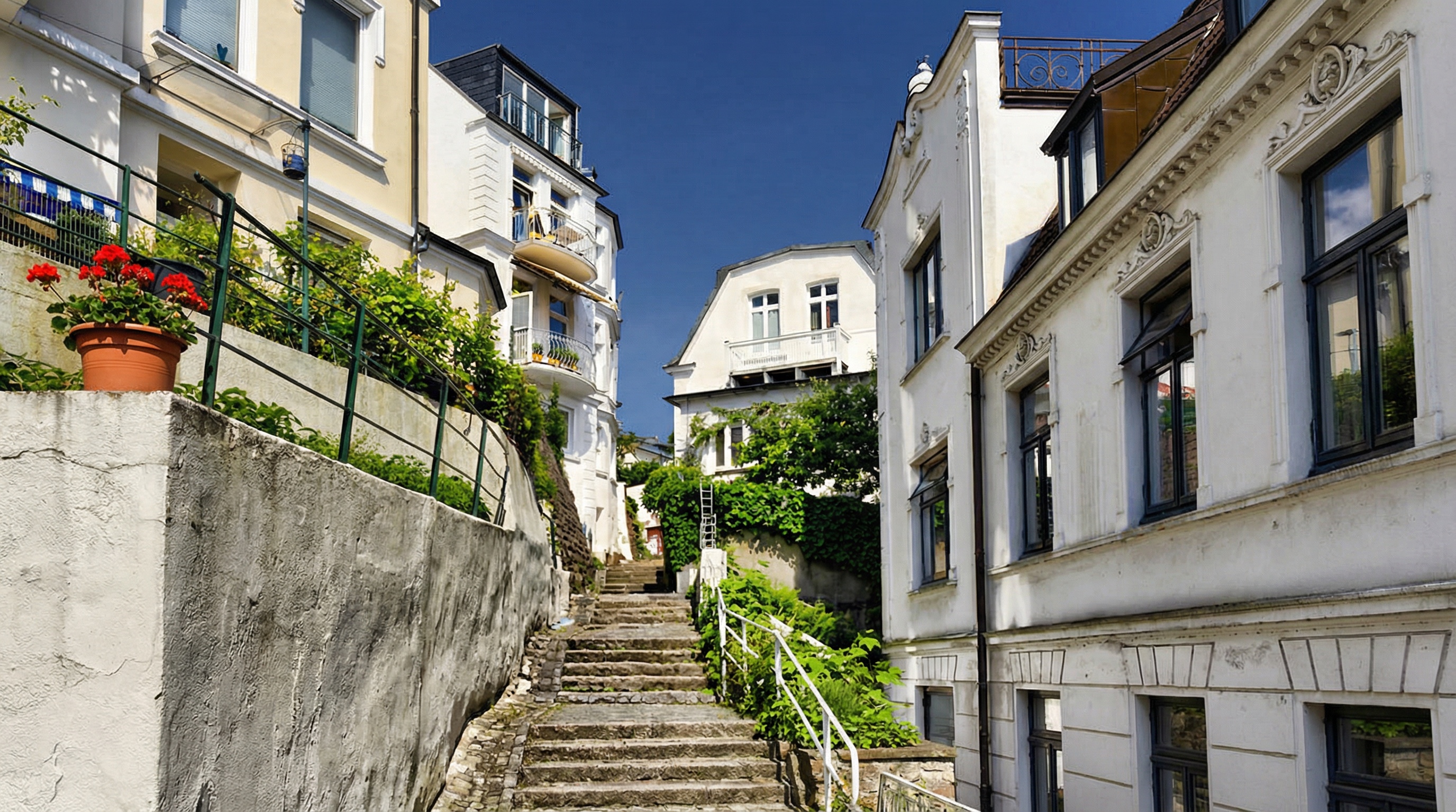 Steile Außentreppe zwischen mehrstöckigen Wohnhäusern mit weißen Fassaden und Balkonen, links Blumentopf mit roten Blumen in Blankenese, Hamburg.