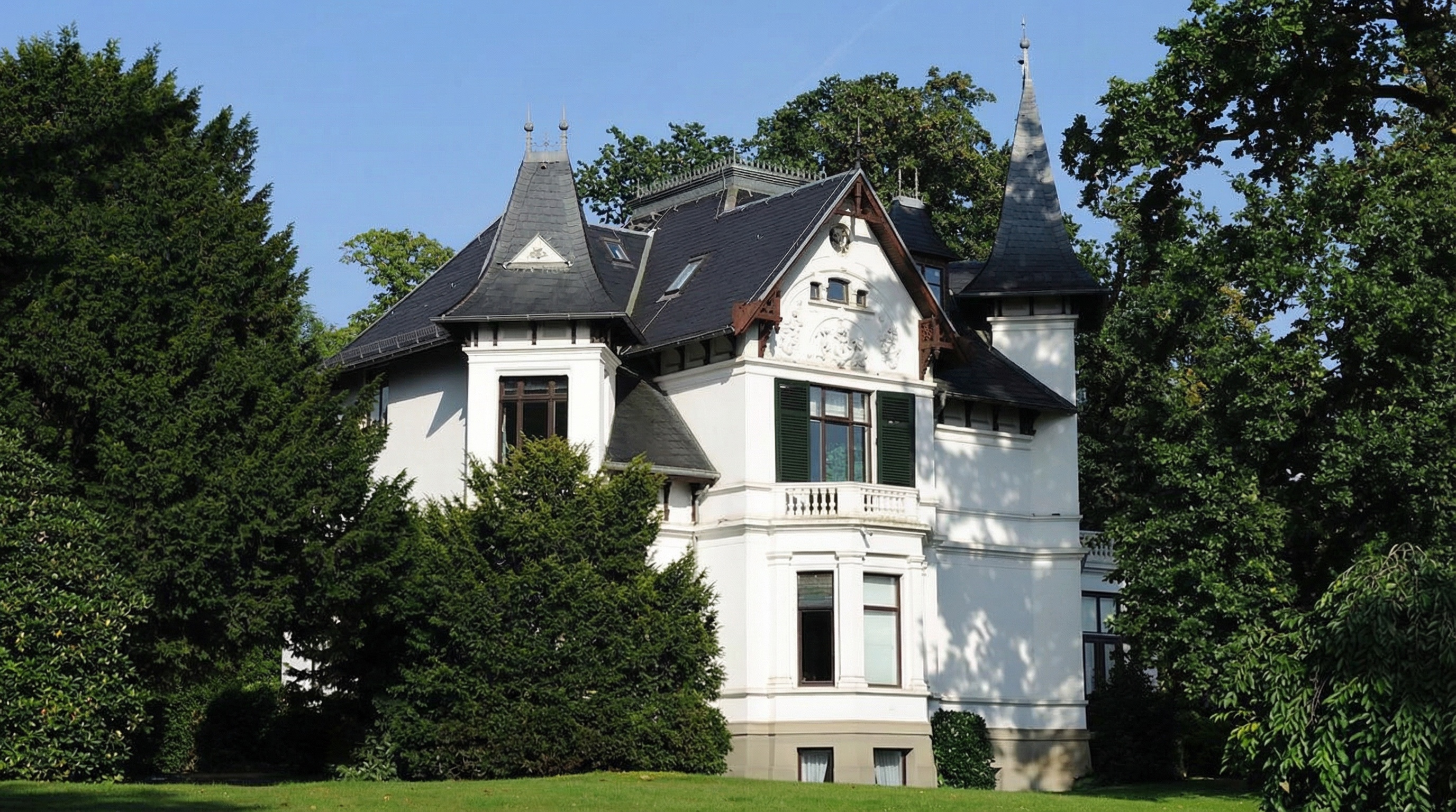 Weißes historisches Gebäude mit spitzen Dächern, grünen Fensterläden und umgeben von Bäumen und Rasen in Nienstedten, Hamburg.
