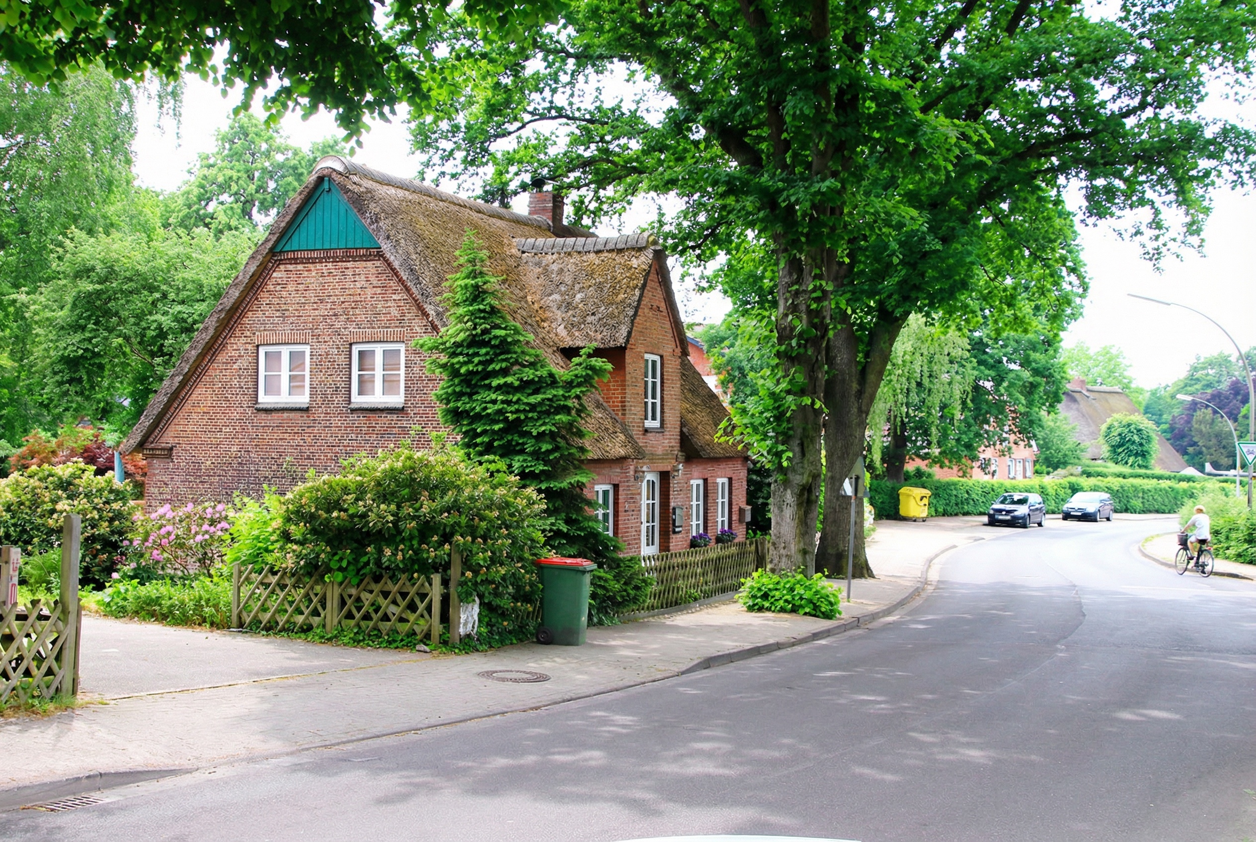 Backsteinhaus mit Reetdach an einer ruhigen Straße, umgeben von Bäumen und Sträuchern, mit Fahrradfahrer auf der Straße in Sülldorf, Hamburg.