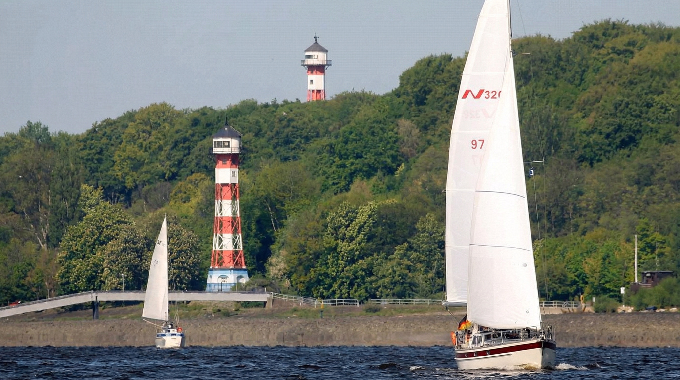 Zwei Segelboote auf dem Wasser vor einem bewaldeten Ufer mit zwei rot-weiß gestreiften Leuchttürmen in Rissen, Hamburg.