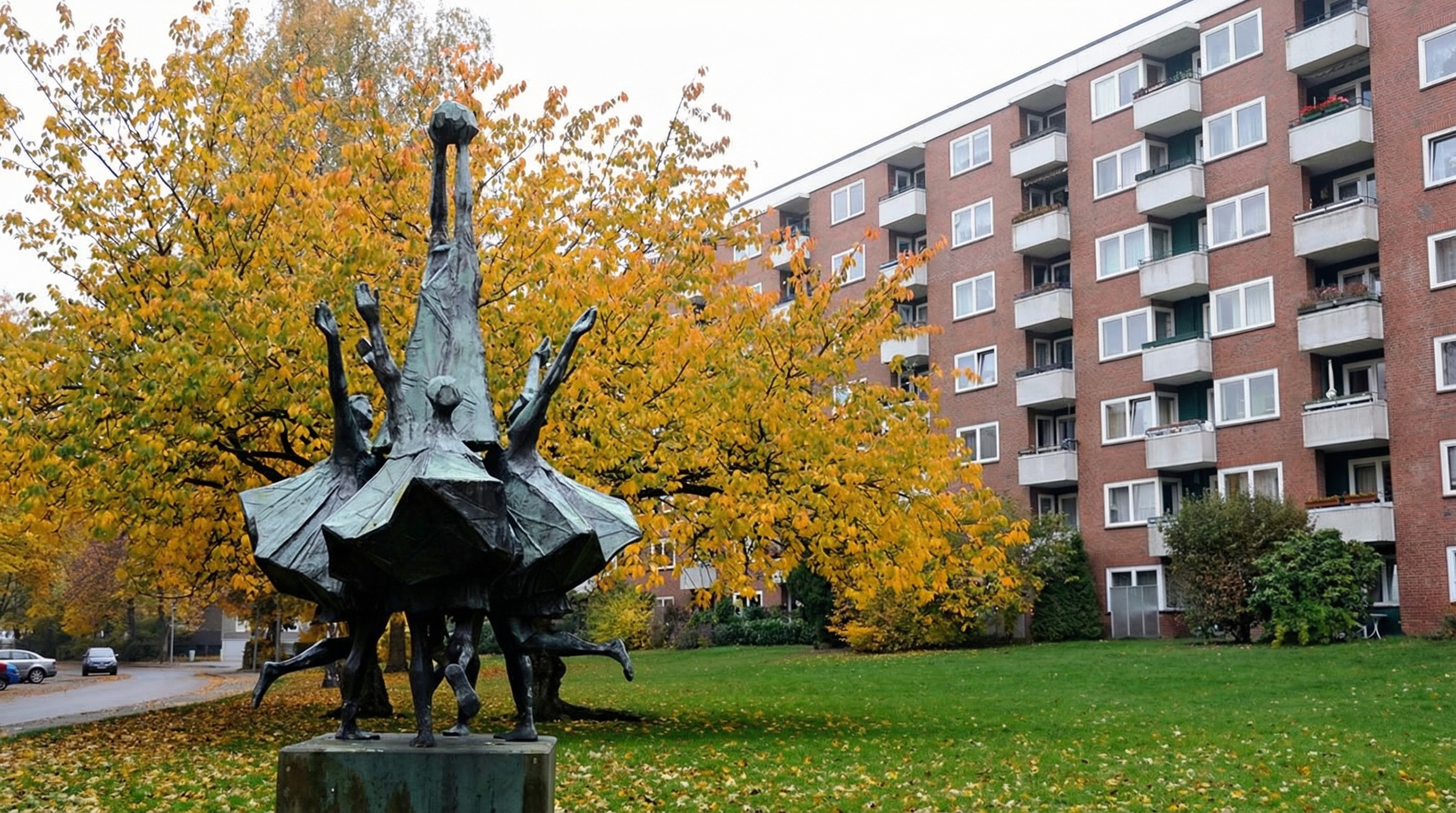 Bronzeskulptur mit mehreren Personen, die sich an den Händen halten und in einem Kreis stehen, im Hintergrund herbstliche Bäume und ein Wohnhaus in Lurup, Hamburg.