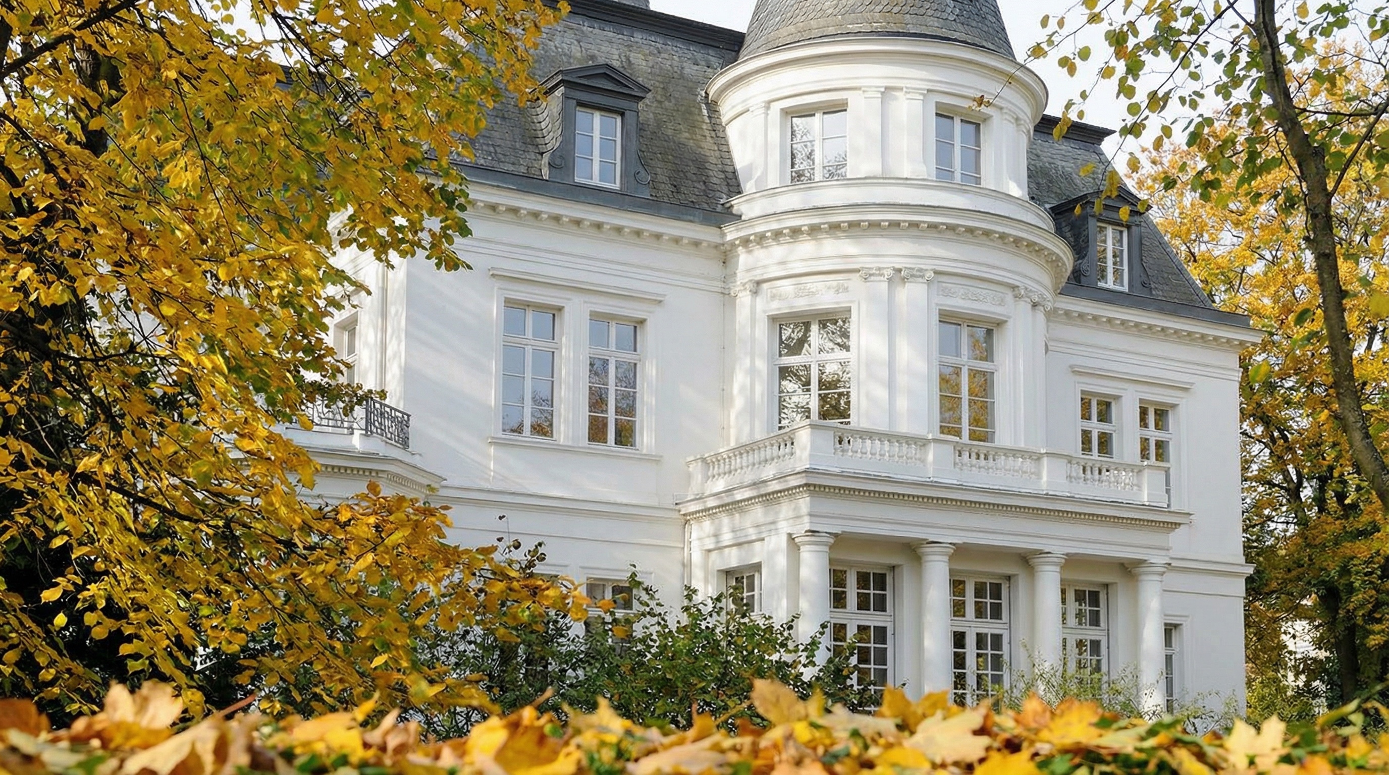 Weißes historisches Gebäude mit Säulen, teilweise von Bäumen mit gelben und grünen Blättern verdeckt in Rotherbaum, Hamburg.
