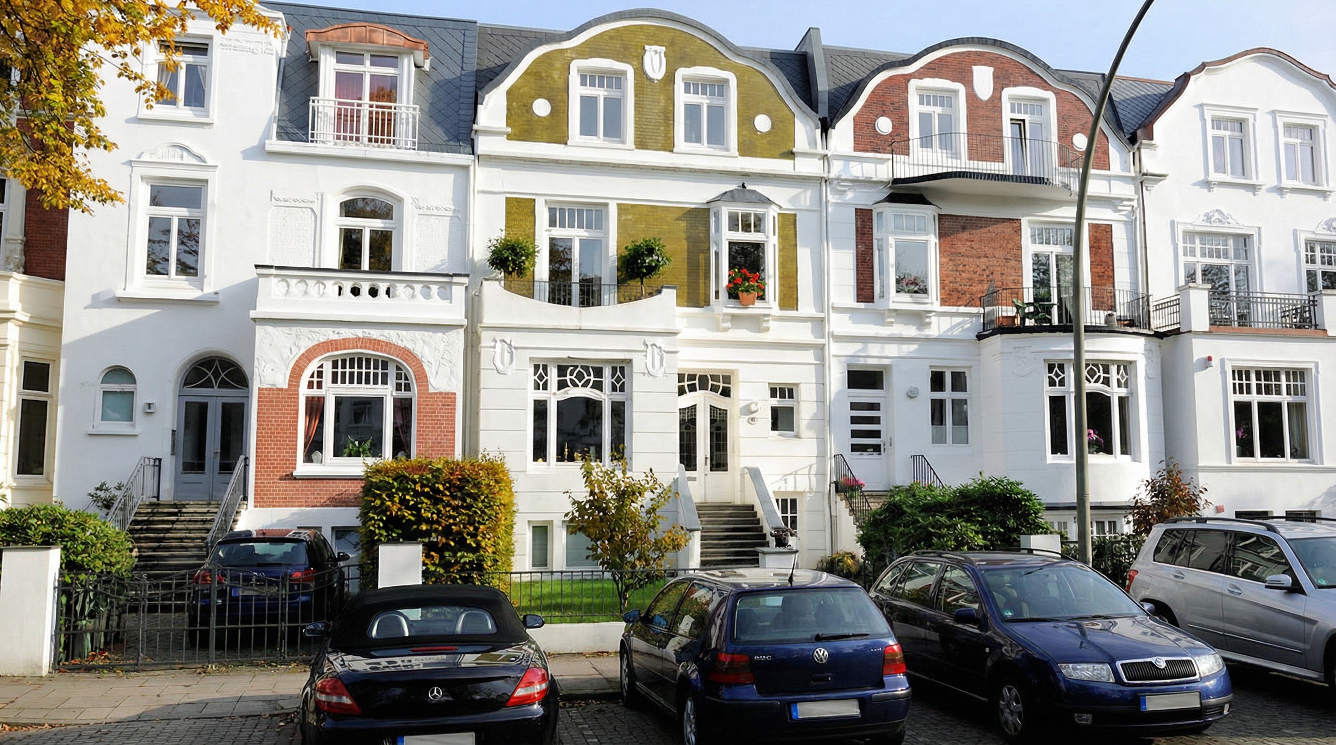 Reihe von fünf mehrstöckigen Stadthäusern mit weißen Fassaden, zwei mit farbigen Giebeln in Rot und Ocker, davor parkende Autos und herbstliche Büsche in Harvestehude, Hamburg.