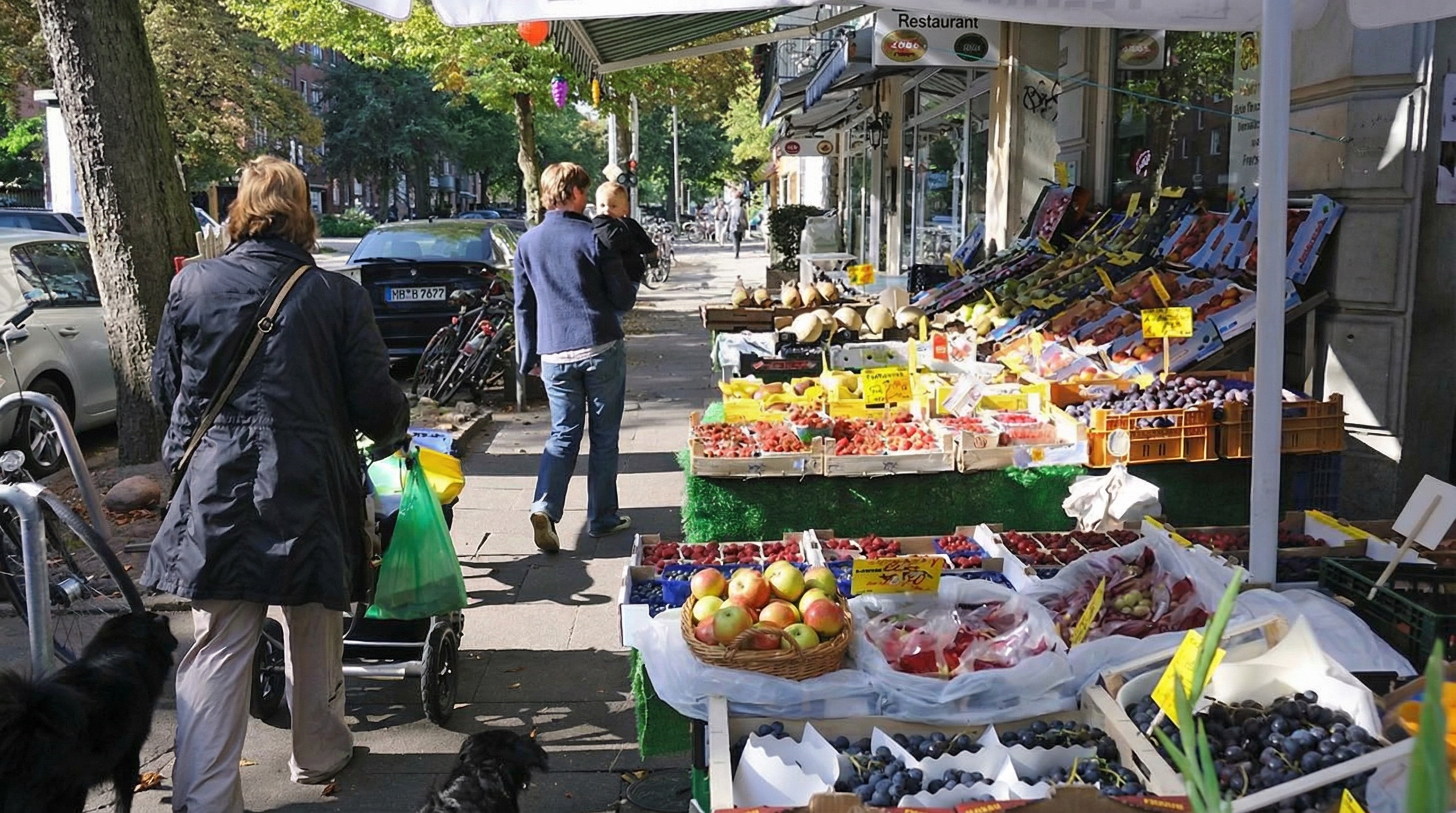 Straßenszene mit mehreren Passanten, einem Hund und einem Marktstand mit Obst und Gemüse unter einem Marktschirm in Hoheluft-West, Hamburg.