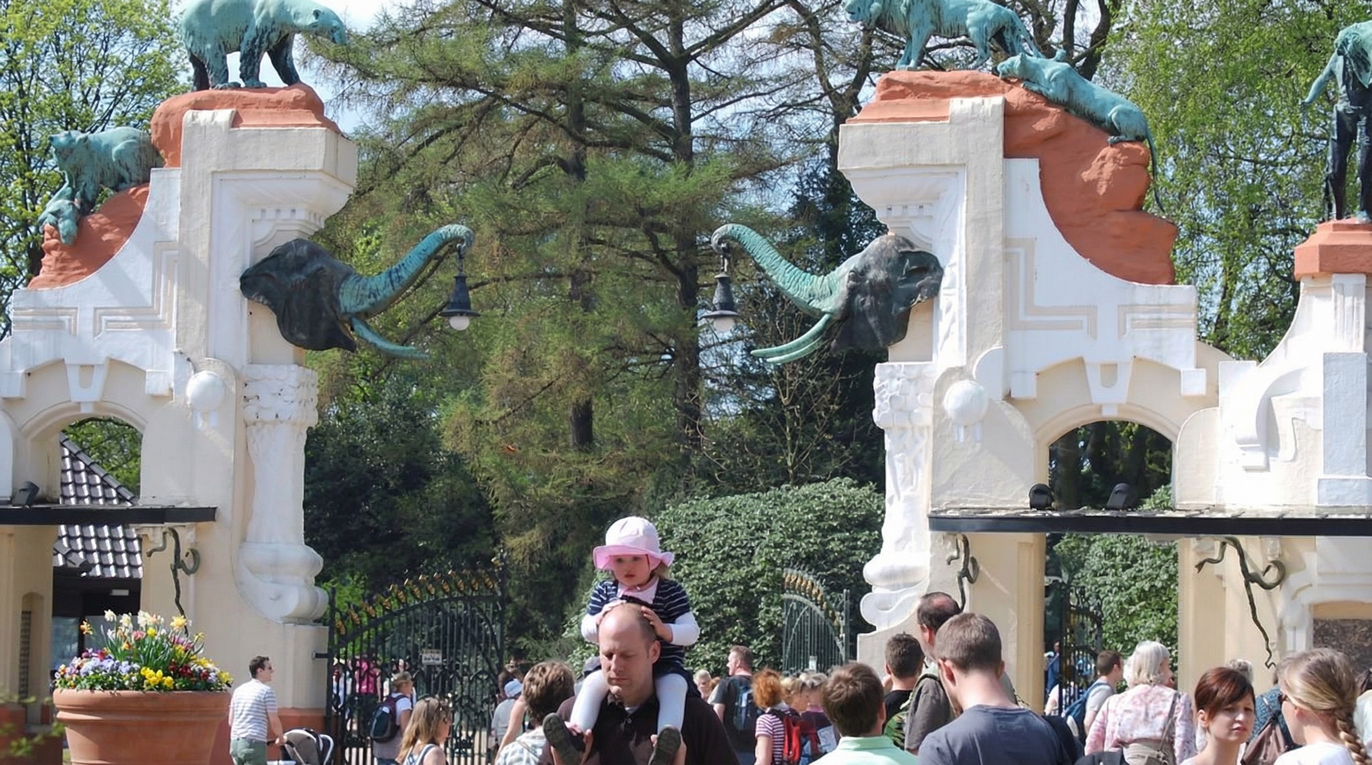 Altes Eingangstor mit zwei großen Elefantenskulpturen des Tierparks Hagenbeck auf weißen Pfeilern, dahinter Bäume und viele Menschen im Freien in Stellingen, Hamburg.