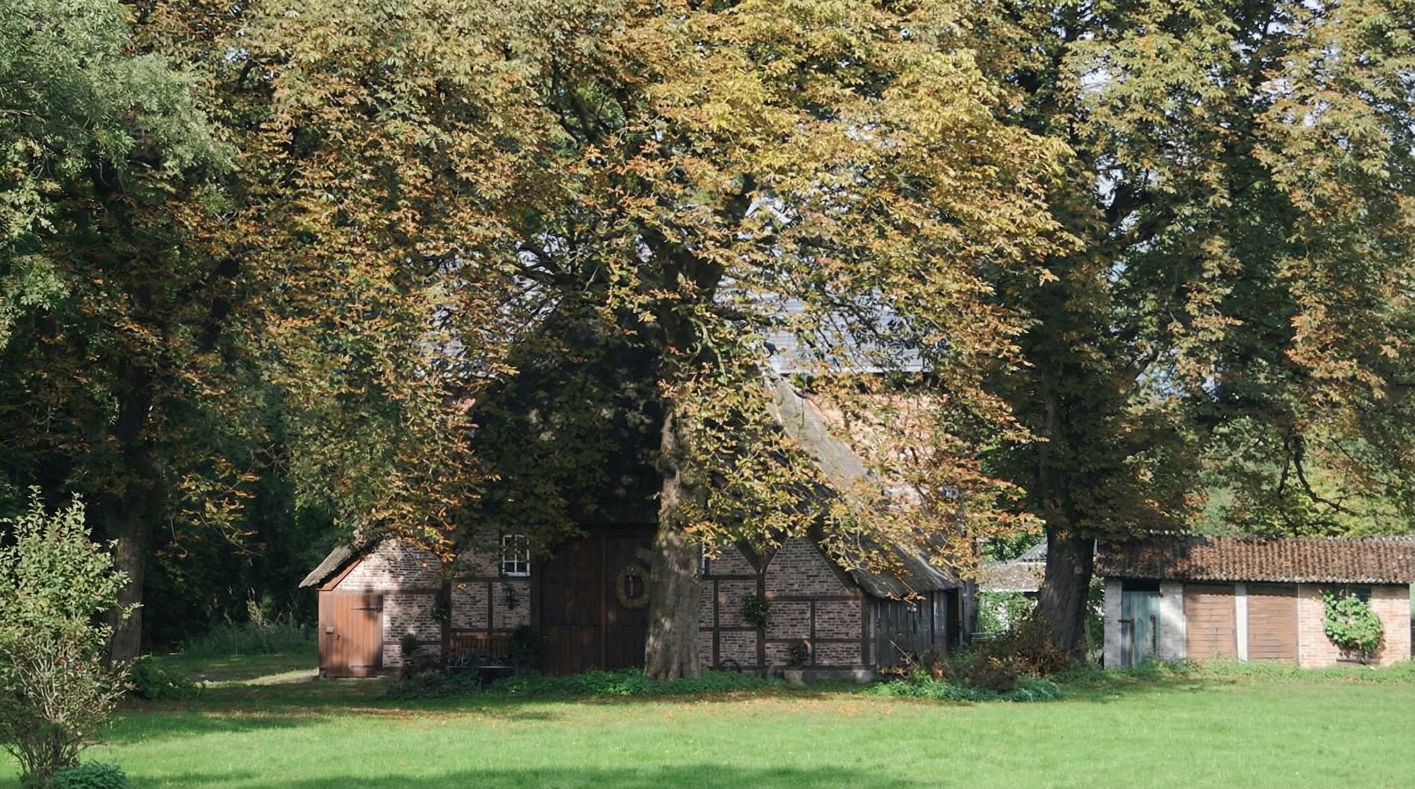 Altes Fachwerkhaus mit mehreren Anbauten, umgeben von großen Bäumen und einer grünen Wiese in Schnelsen, Hamburg.