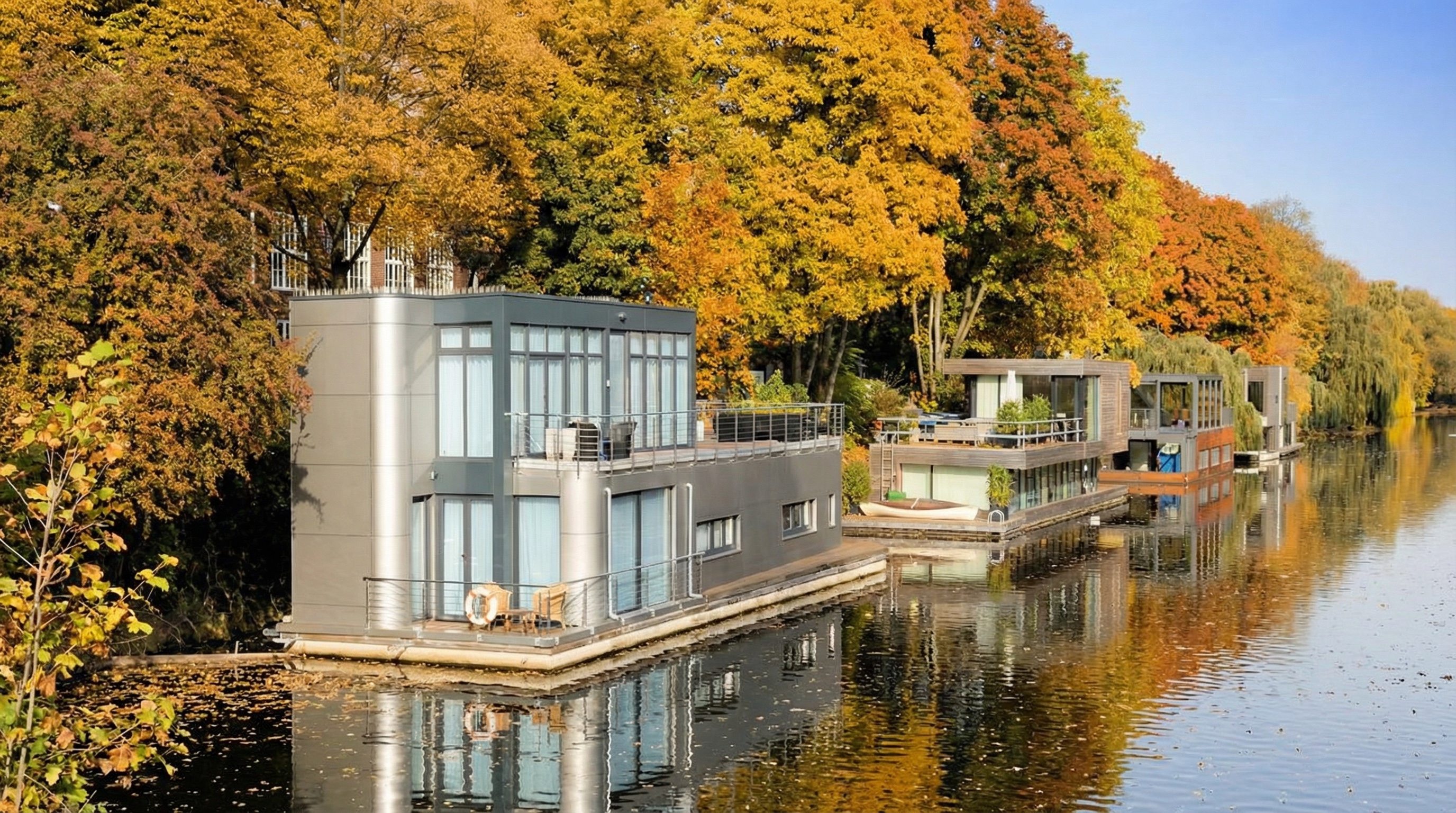 Zwei moderne Hausboote am Ufer eines Kanals, umgeben von herbstlich gefärbten Bäumen unter blauem Himmel in Barmbek-Süd, Hamburg.
