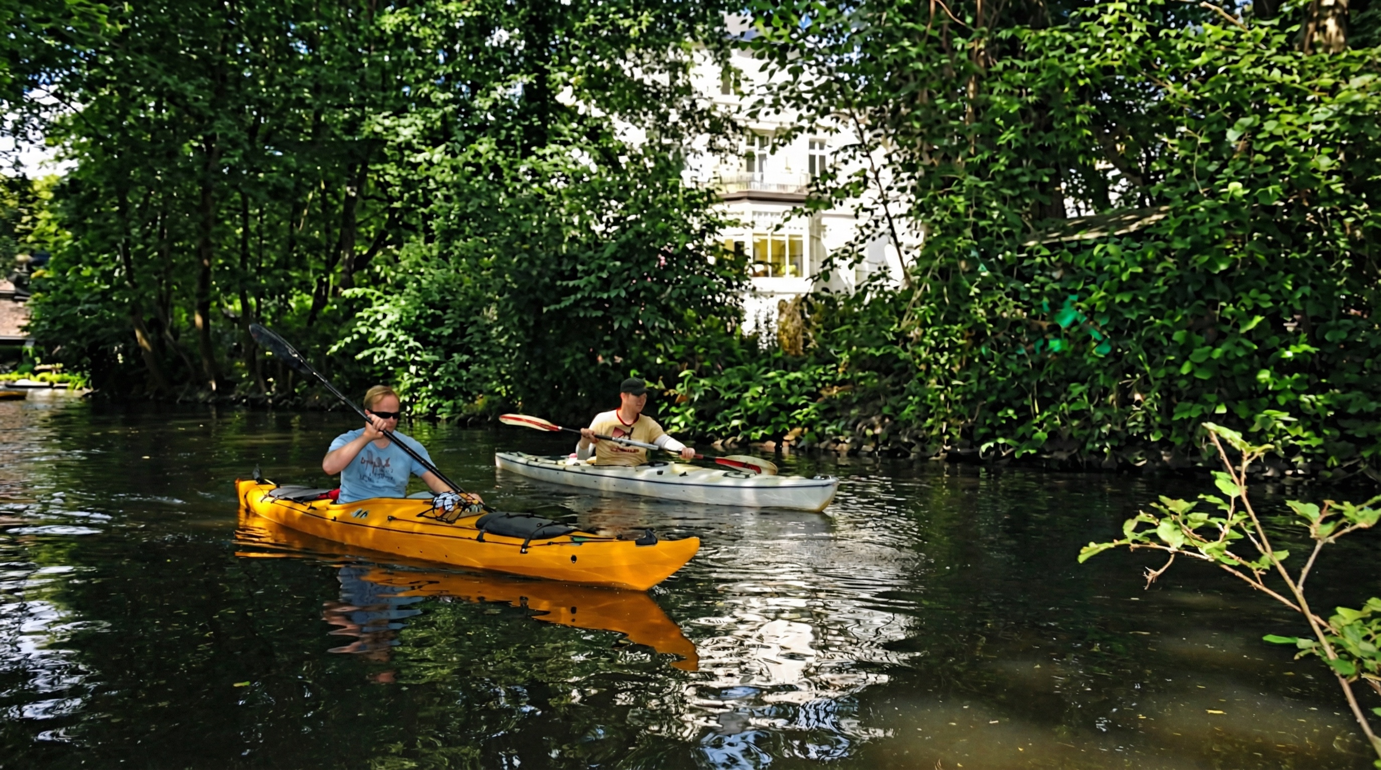 Zwei Personen paddeln in einem gelben und einem weißen Kajak auf einem von Bäumen gesäumten Gewässer in Winterhude, Hamburg.