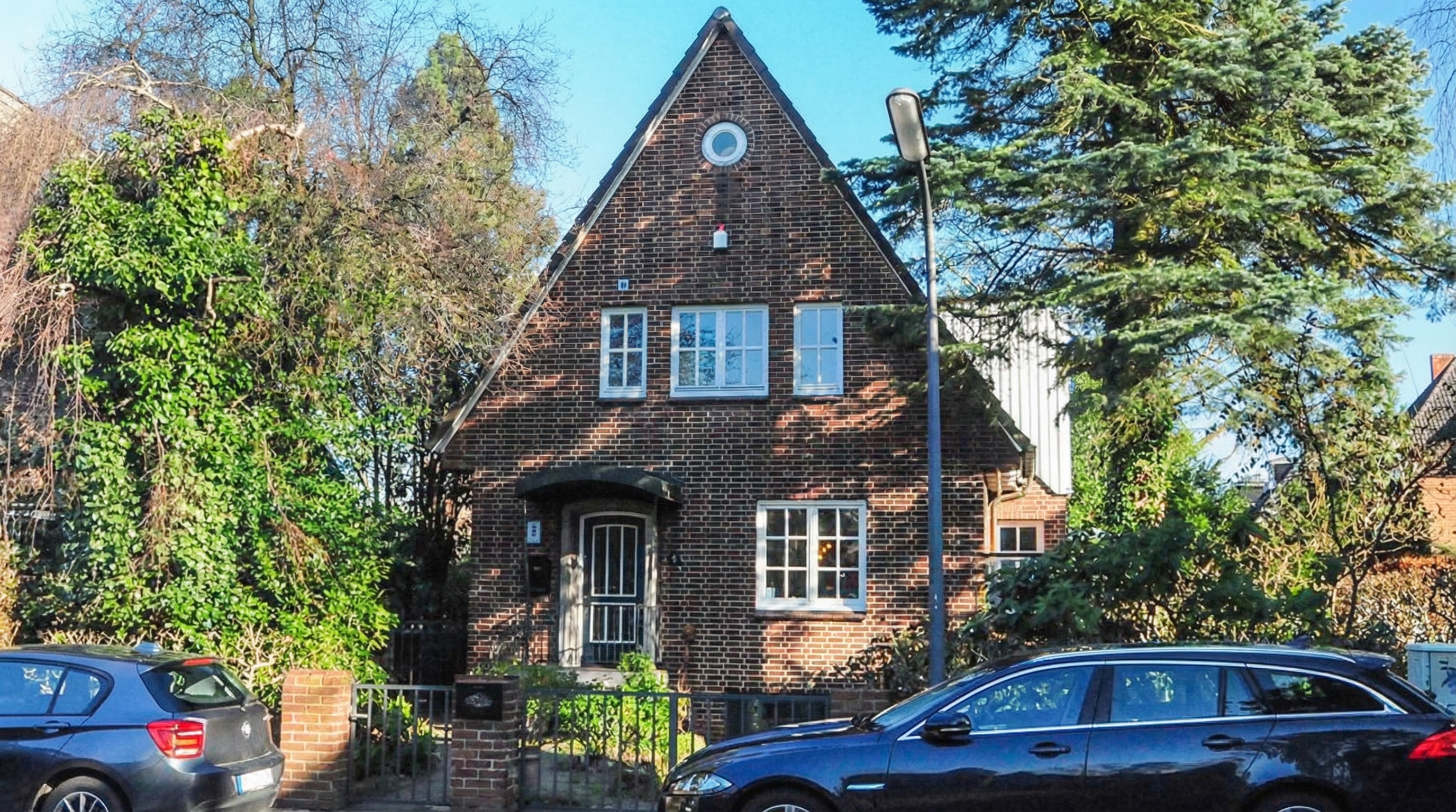 Zweistöckiges, dreieckiges Backsteinhaus mit weißen Fensterrahmen, umgeben von Bäumen und parkenden Autos davor in Alsterdorf, Hamburg.