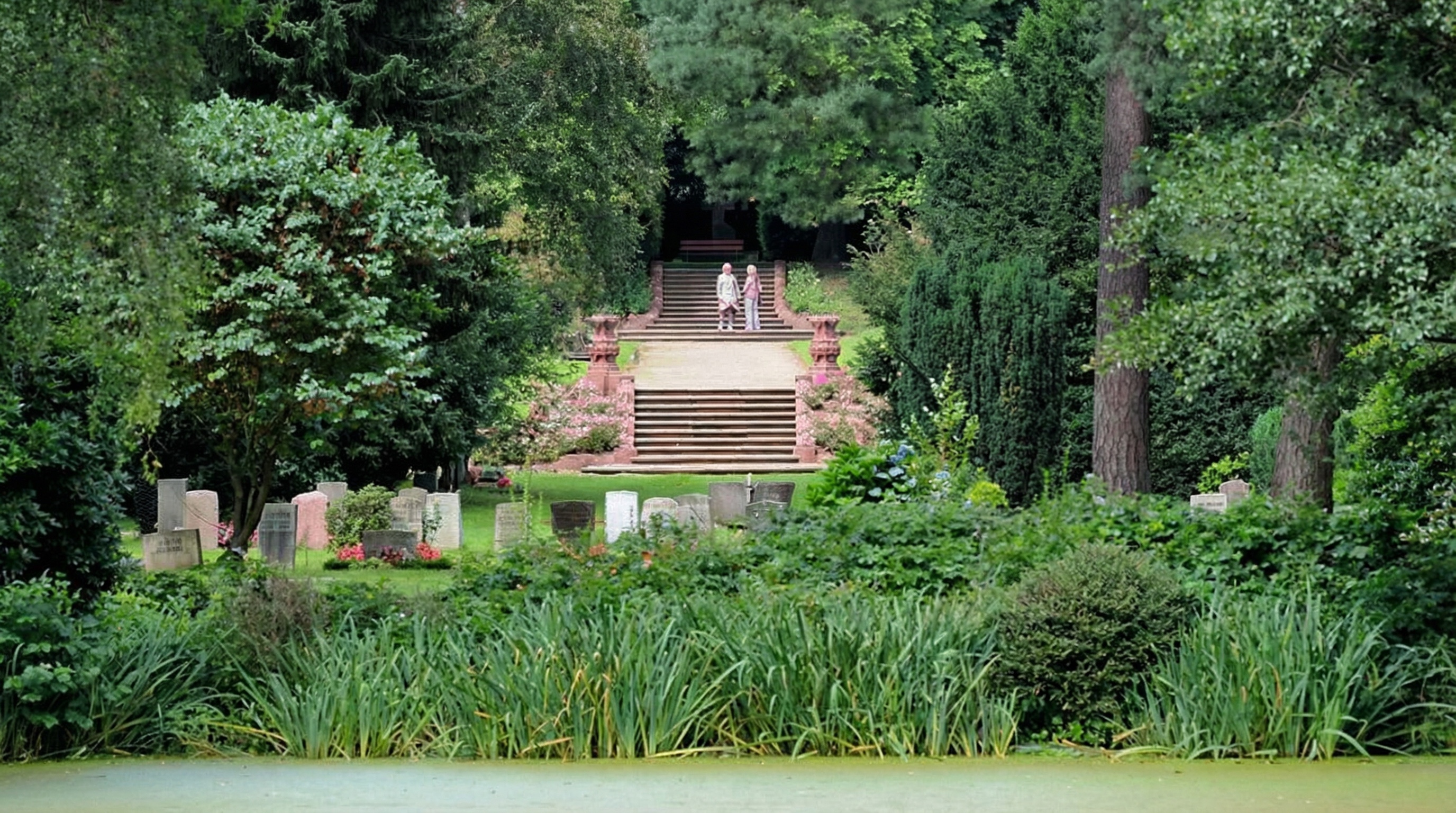 Park mit Teich im Vordergrund, dahinter Treppenaufgang und zwei Personen auf einer Plattform, umgeben von Bäumen und Grabsteinen auf dem Friedhof in Ohlsdorf, Hamburg.
