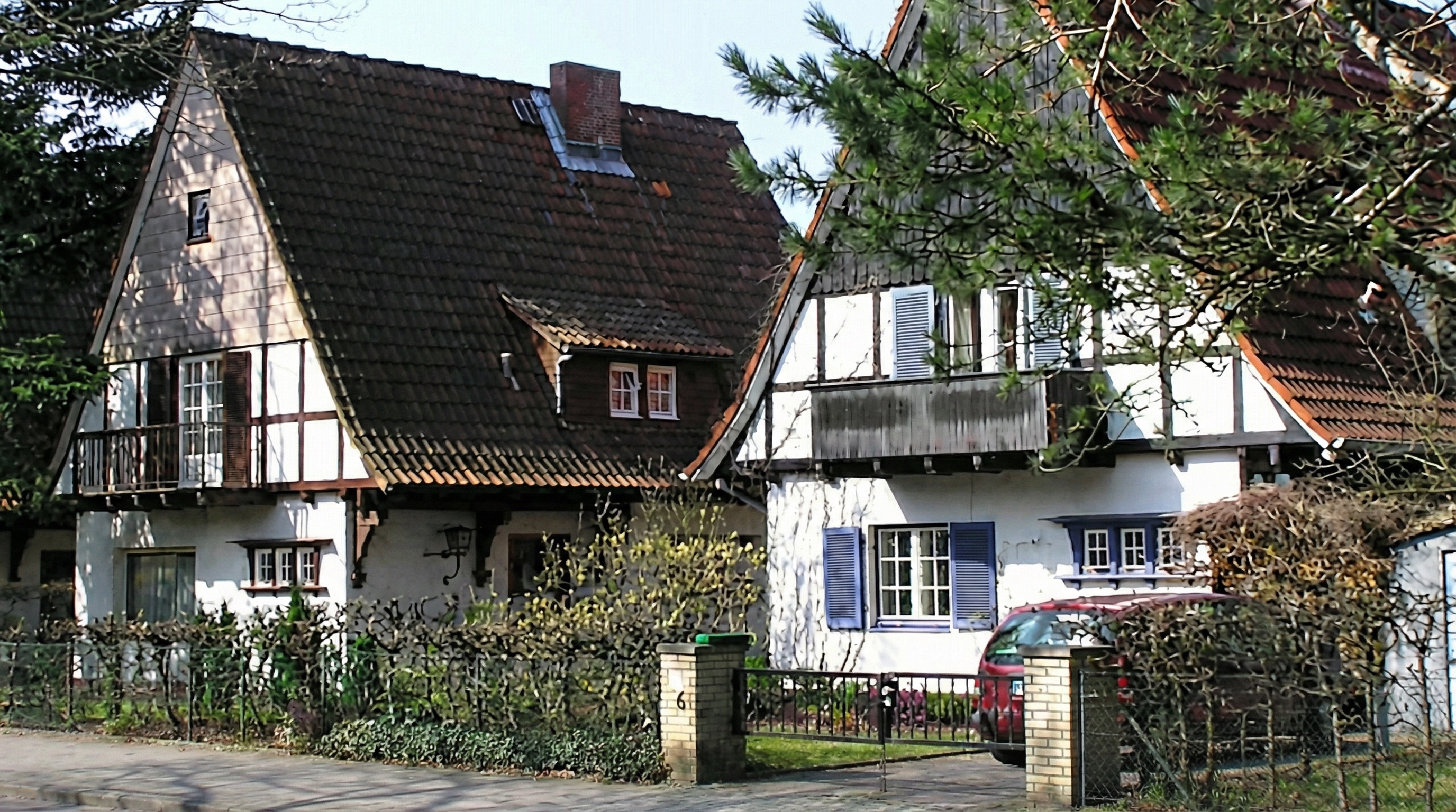 Zwei Fachwerkhäuser mit braunen Dächern, umgeben von Büschen und einem Zaun mit Tor, davor ein rotes Auto in Langenhorn, Hamburg.