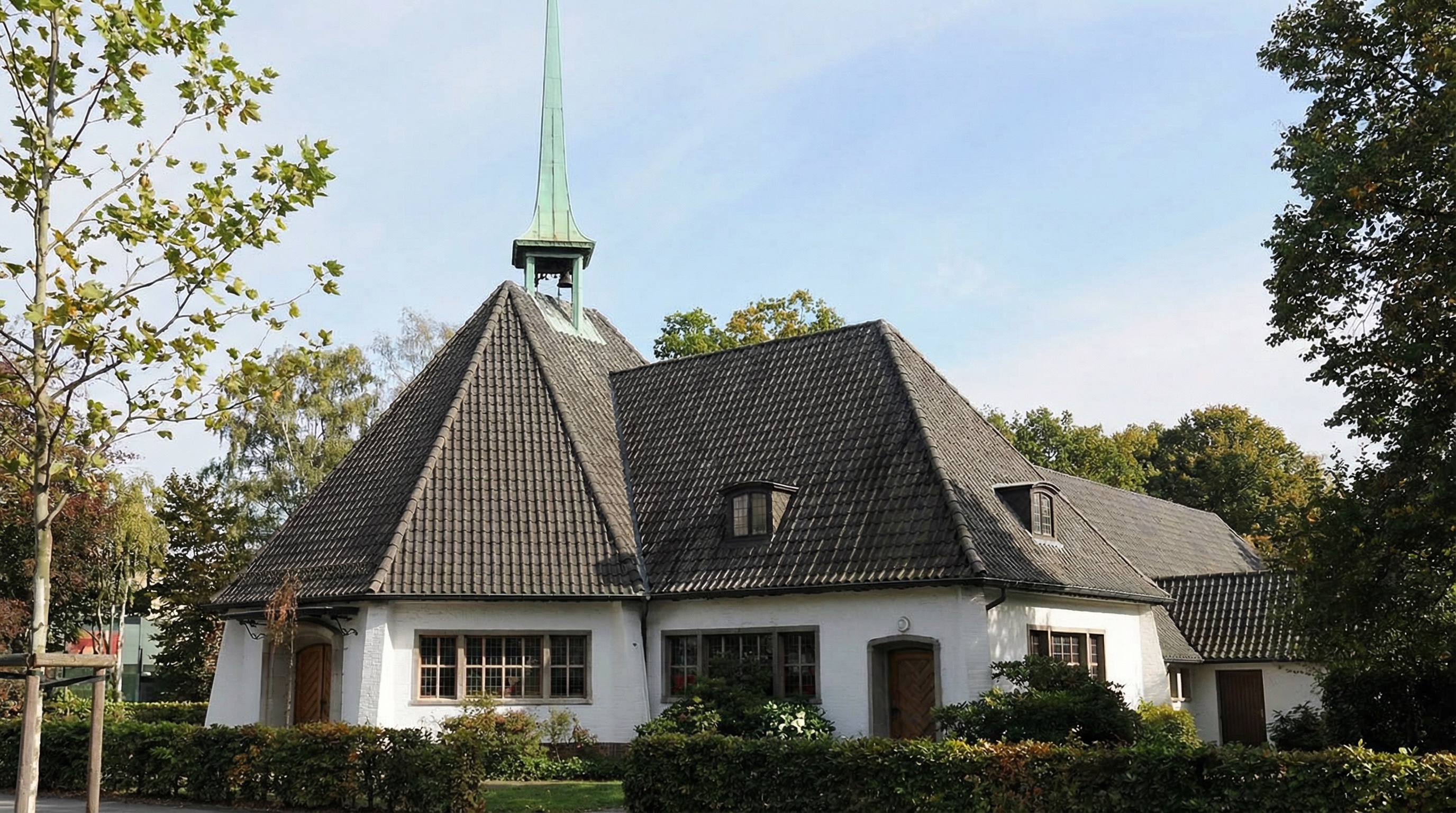Weißes Kirchengebäude mit dunklem Satteldach und grünem Turmspitz, umgeben von Bäumen und Hecken in Tonndorf, Hamburg.