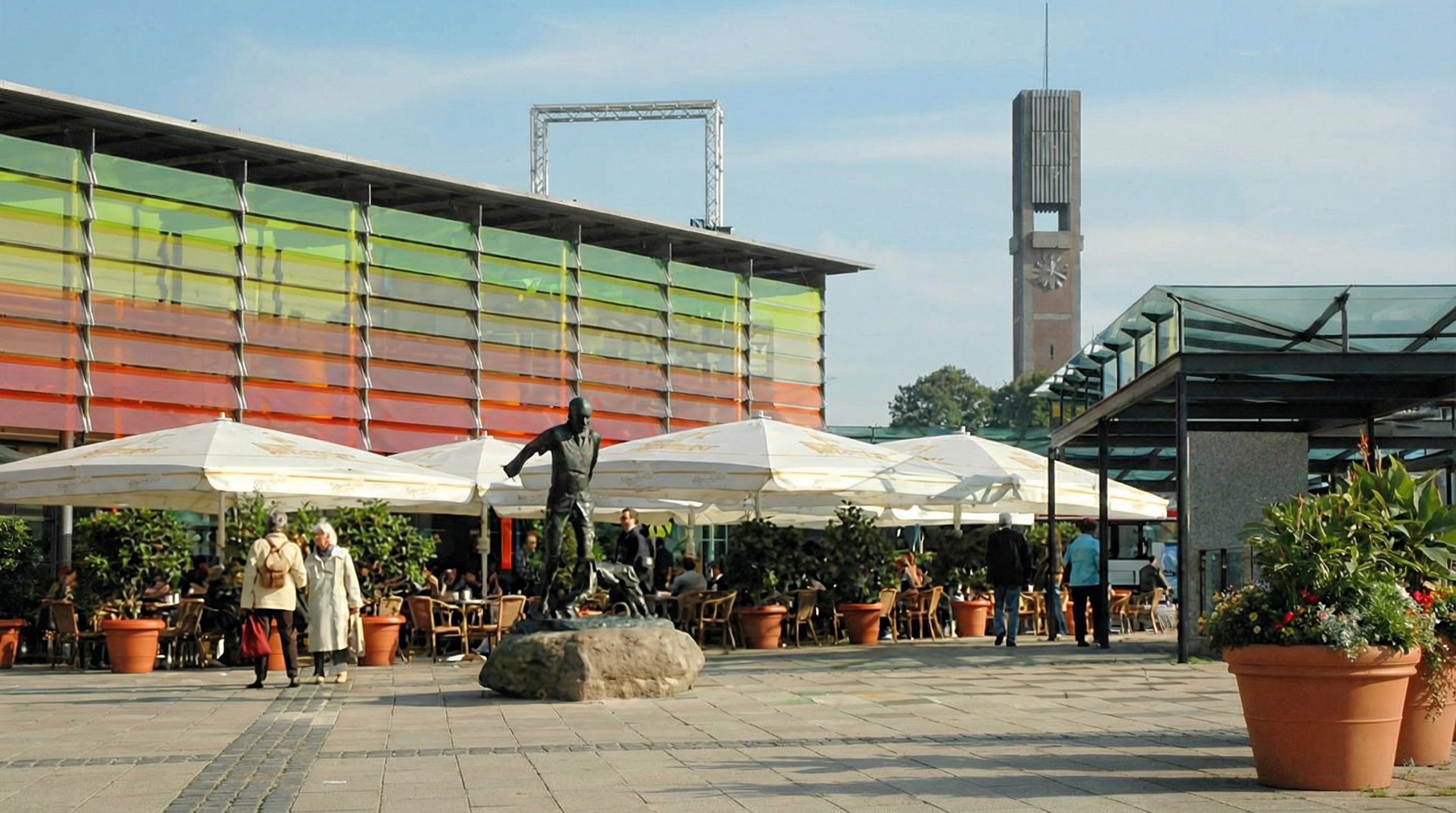 Platz mit bronzener Statue auf Felsen, mehreren großen Sonnenschirmen und buntem Gebäude mit Restaurant im Hintergrund in Wandsbek, Hamburg.