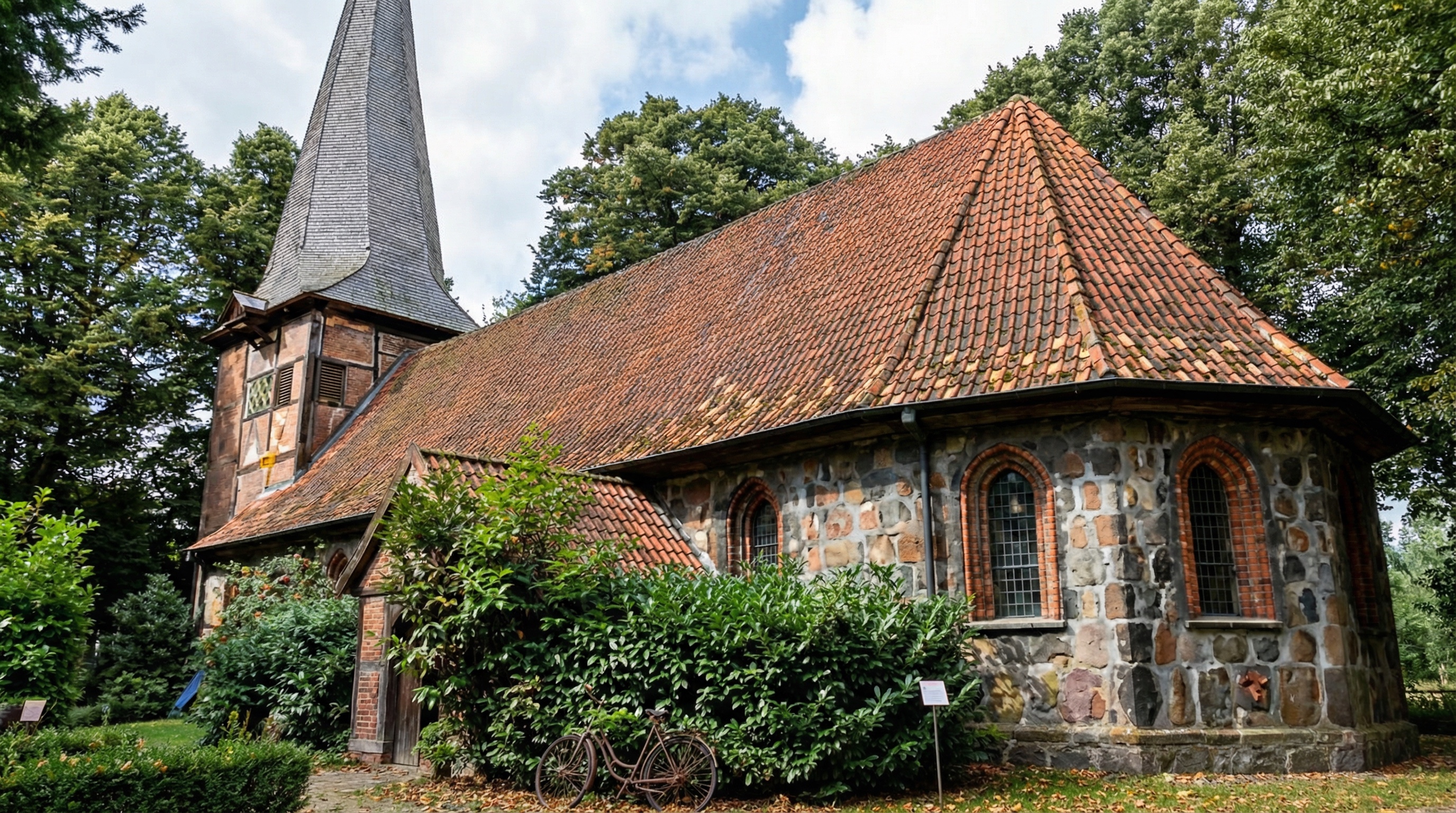 Kirche aus Feldsteinen mit rotem Ziegeldach und spitzem Turm, umgeben von Bäumen und Sträuchern in Rahlstedt, Hamburg.